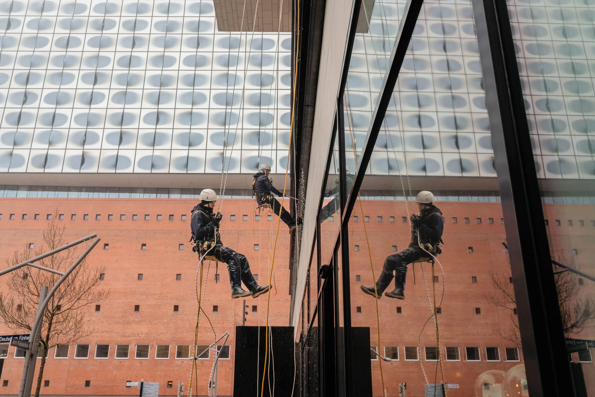 Two workers in safety gear cleaning the exterior glass of a building, with their reflection visible in the glass.
