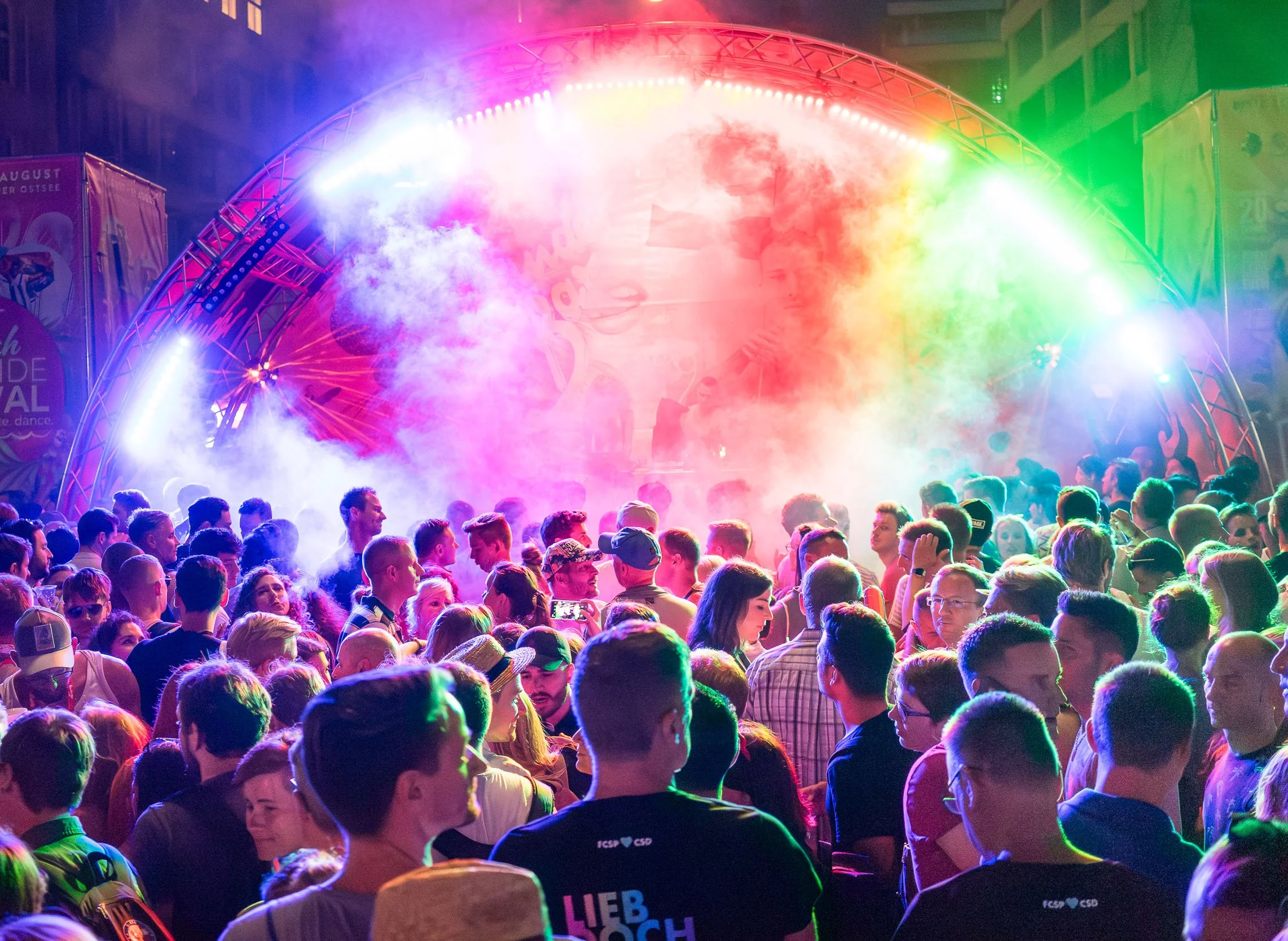 Crowd of people dancing and enjoying a concert or festival with colorful stage lights and smoke effects.