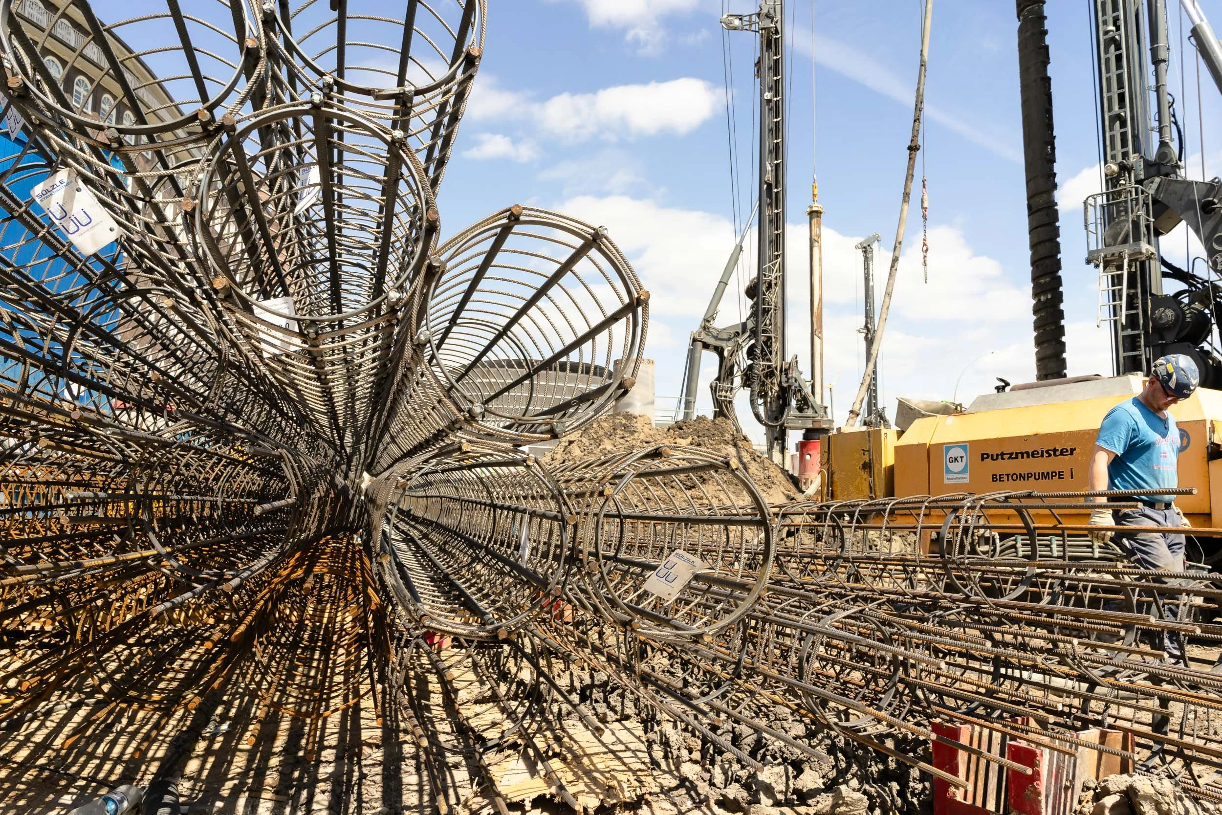 Construction site with steel rebar and large machinery, worker in blue shirt and safety helmet near yellow concrete pump labeled 'Putzmeister'
