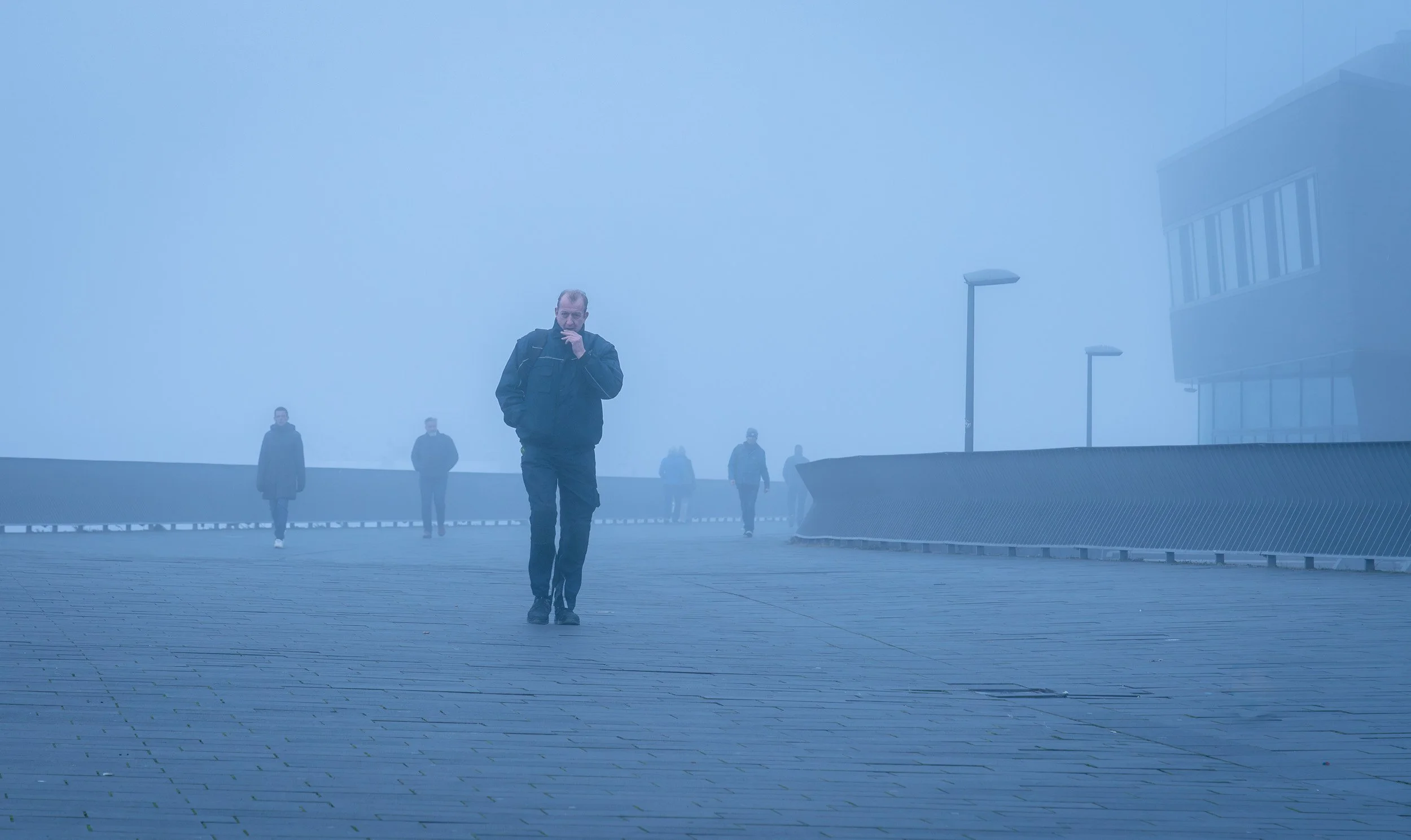 Man walking on a foggy outdoor walkway with other people in the background and modern buildings around.