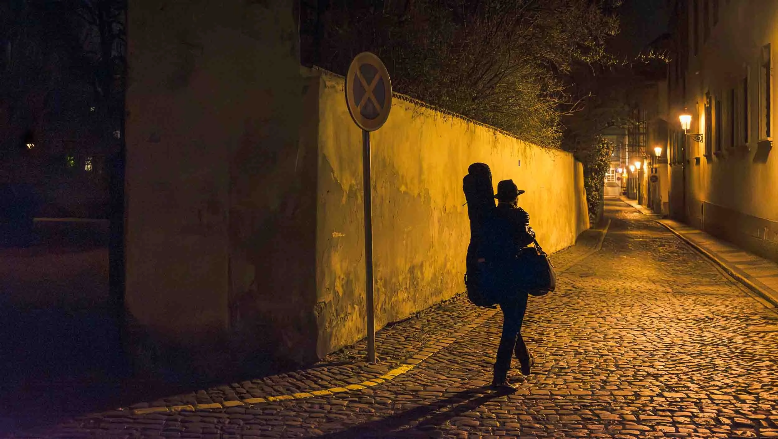A person walking on a cobblestone street at night with street lights illuminating the area. The person is wearing a hat and carrying a bag, walking past a yellow wall with a no parking sign.