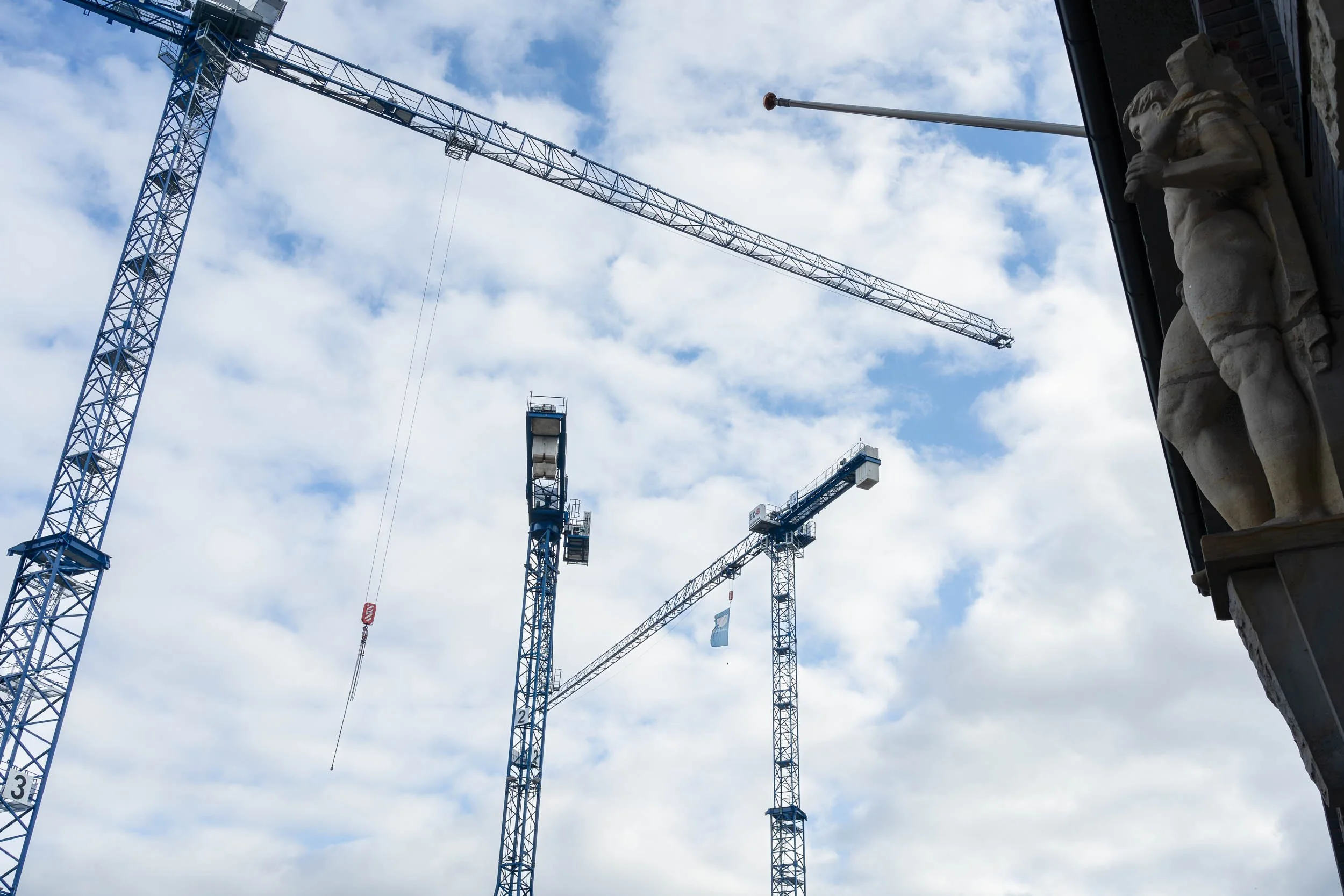 Multiple blue construction cranes against a partly cloudy sky, with a stone sculpture of a man holding a sword on the right side.