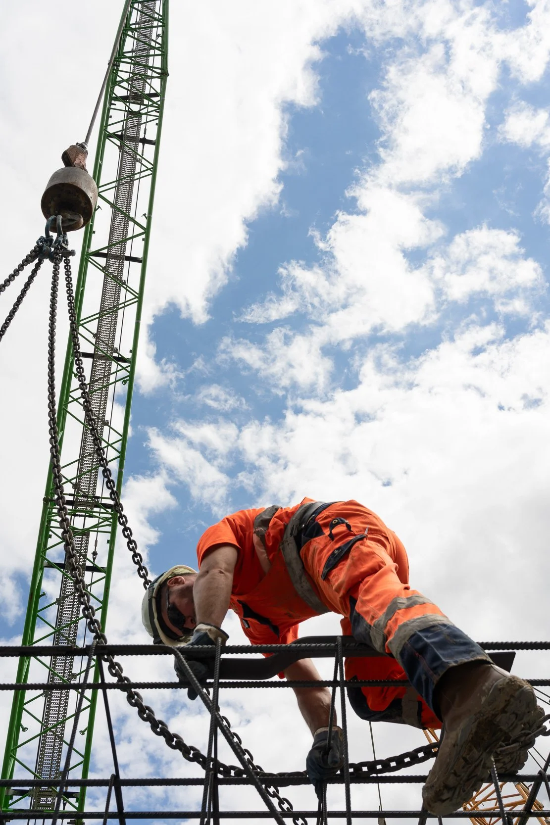 Construction worker wearing an orange safety suit and helmet, climbing on rebar at a construction site with a green crane and partly cloudy sky in the background.