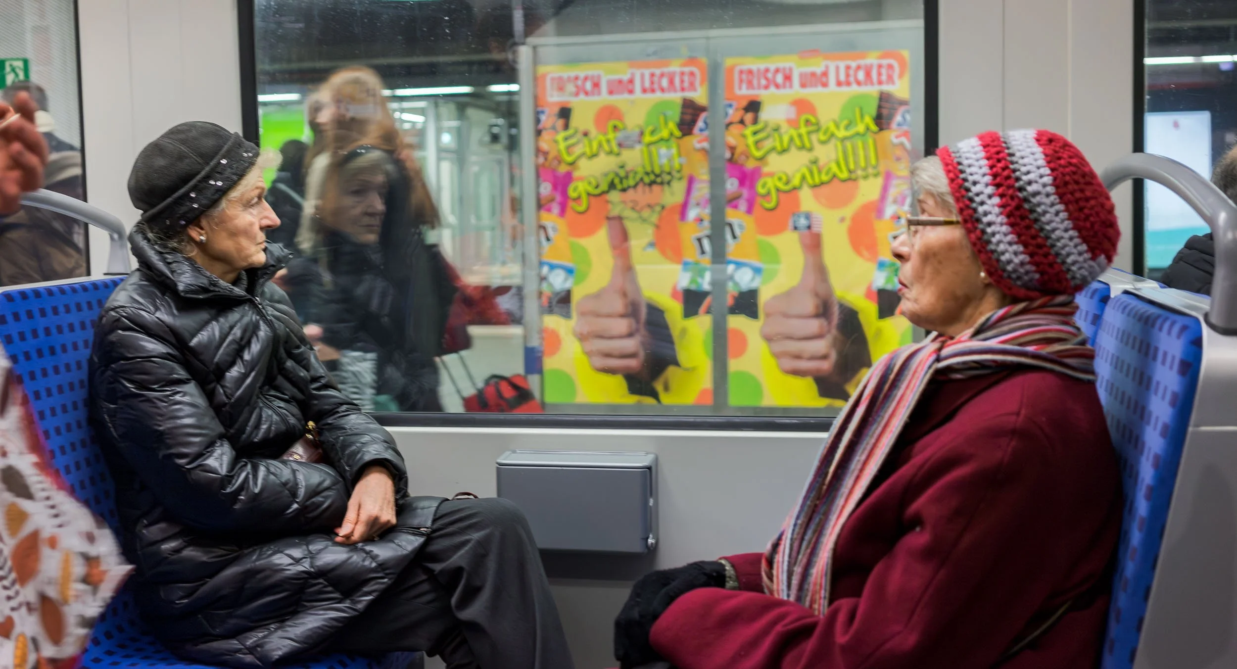 Two older women sitting across from each other on a train, wearing winter clothing and hats, with a window behind them showing a colorful advertisement and some reflections of other passengers.