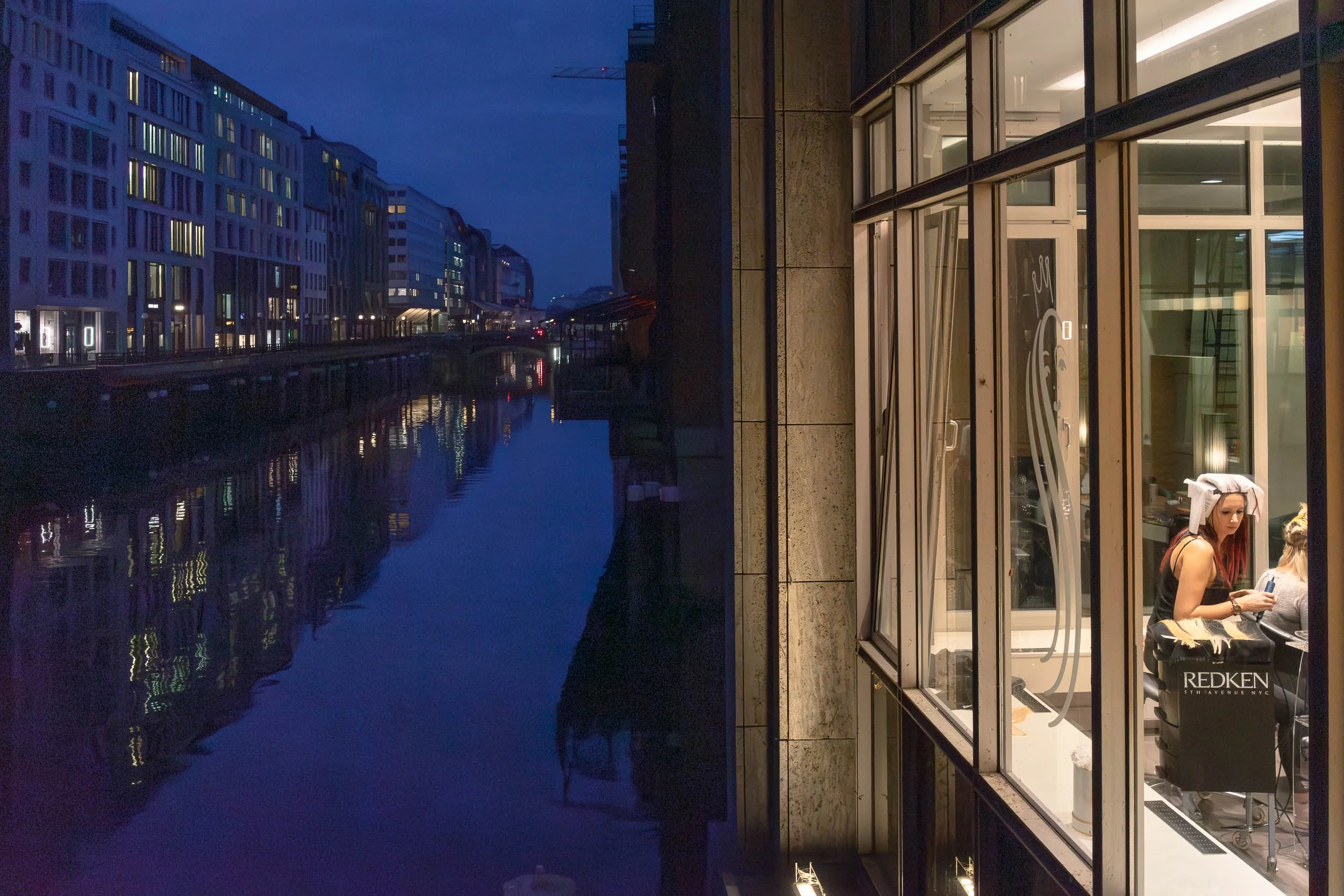 Night view of a city canal with modern buildings on the opposite side. Inside a hair salon, a woman with hair rollers is sitting on a chair getting her hair done, with a Redken sign visible.