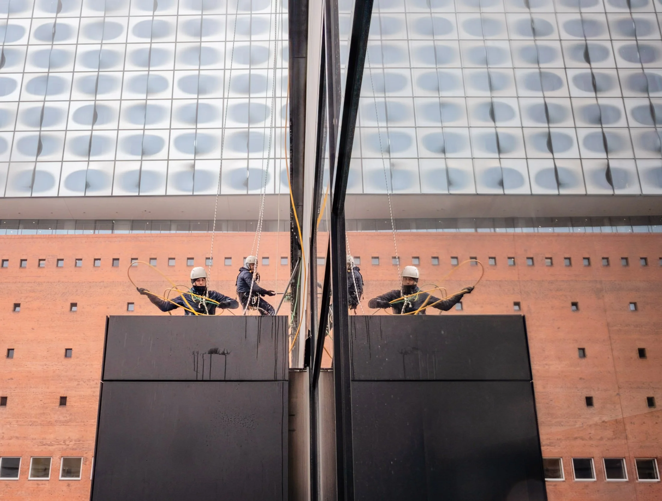 Two workers wearing safety helmets and gloves cleaning the exterior of a glass building with squeegees, reflected in the glass.