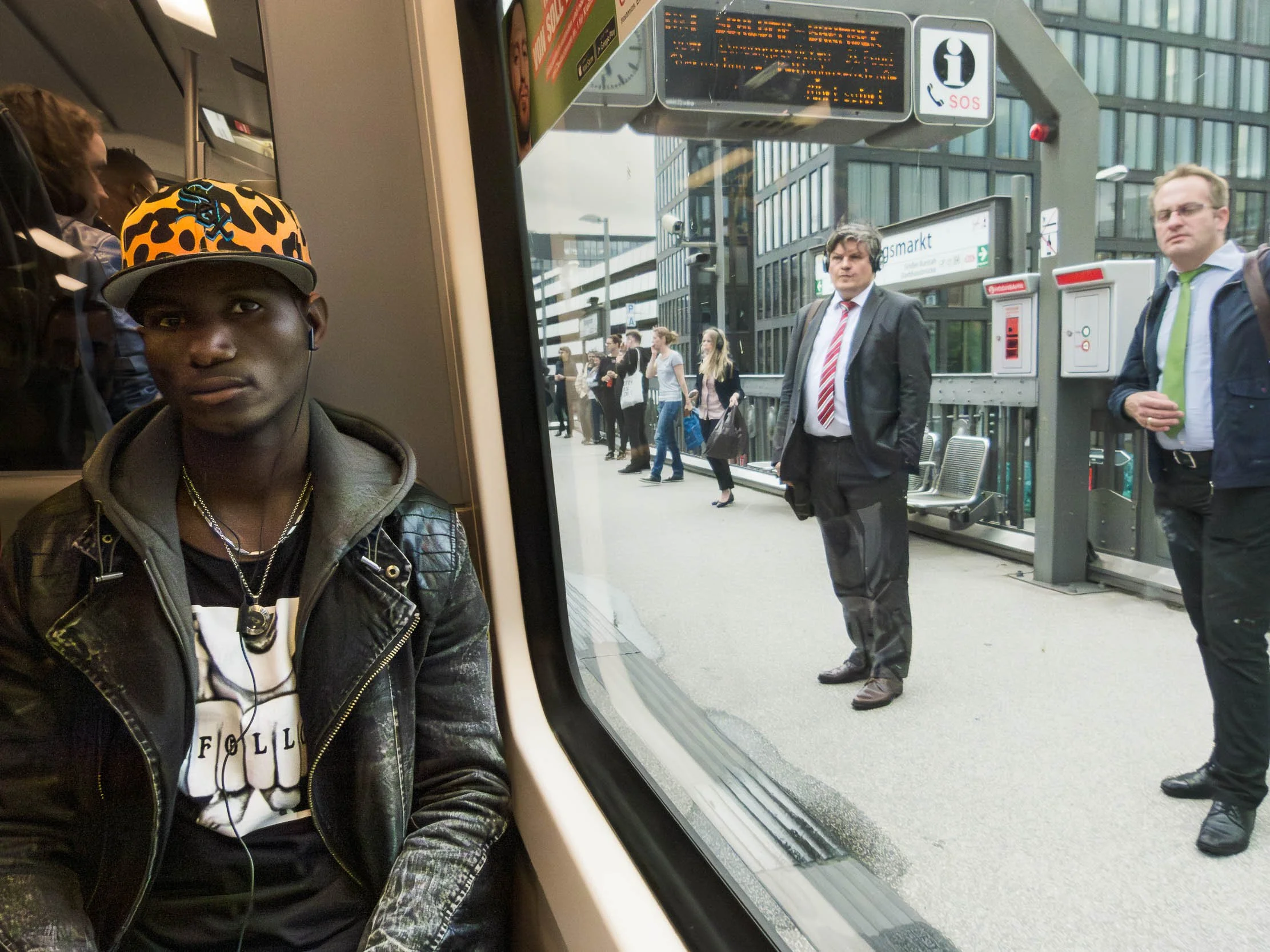 A young man with dark skin wearing a leopard print baseball cap, a leather jacket, and earphones, sitting inside a train or subway, with a bustling city train station visible through the window behind him.