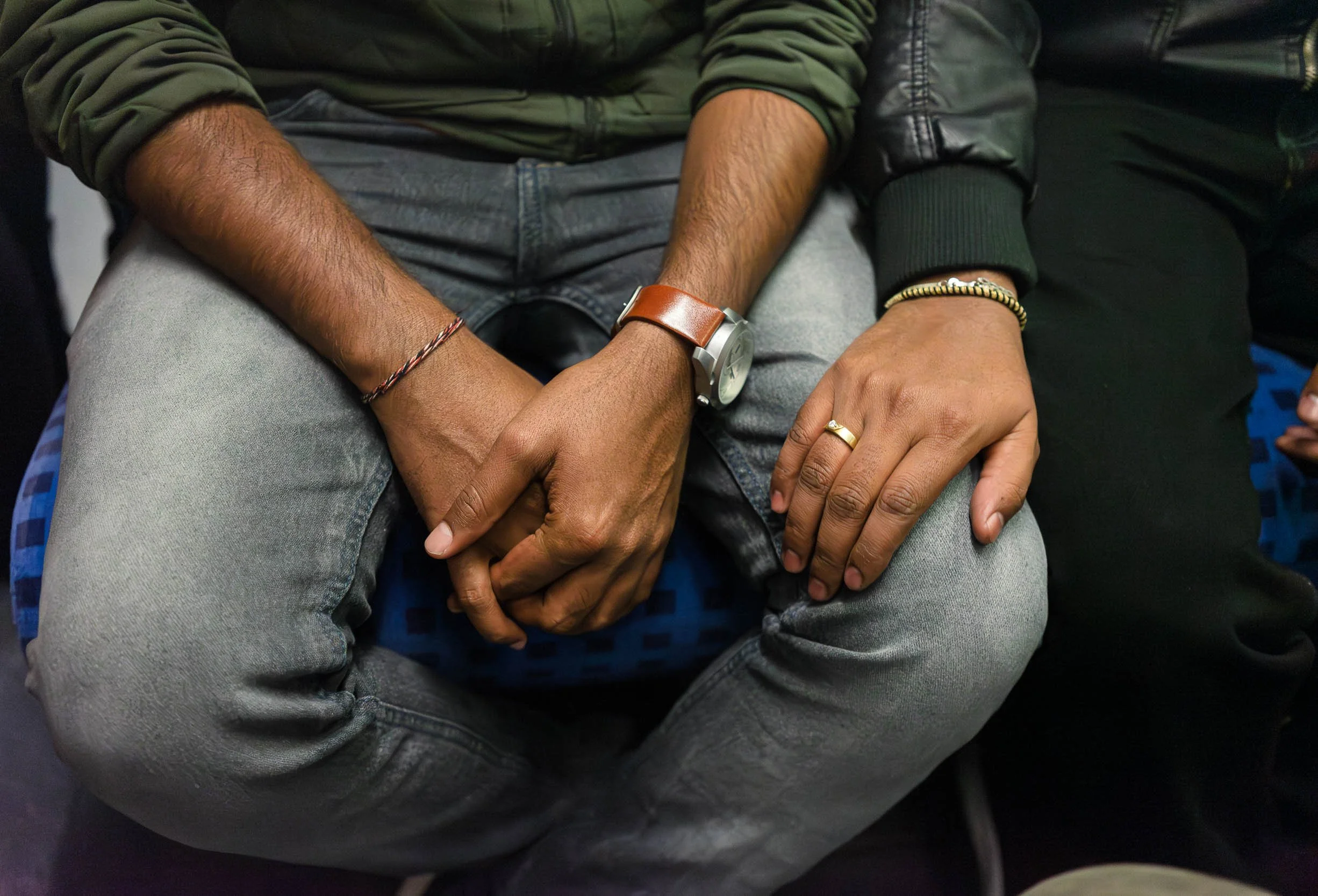 Close-up of two people sitting side by side with their hands resting on their laps, wearing casual clothing and accessories such as watches, rings, and bracelets.