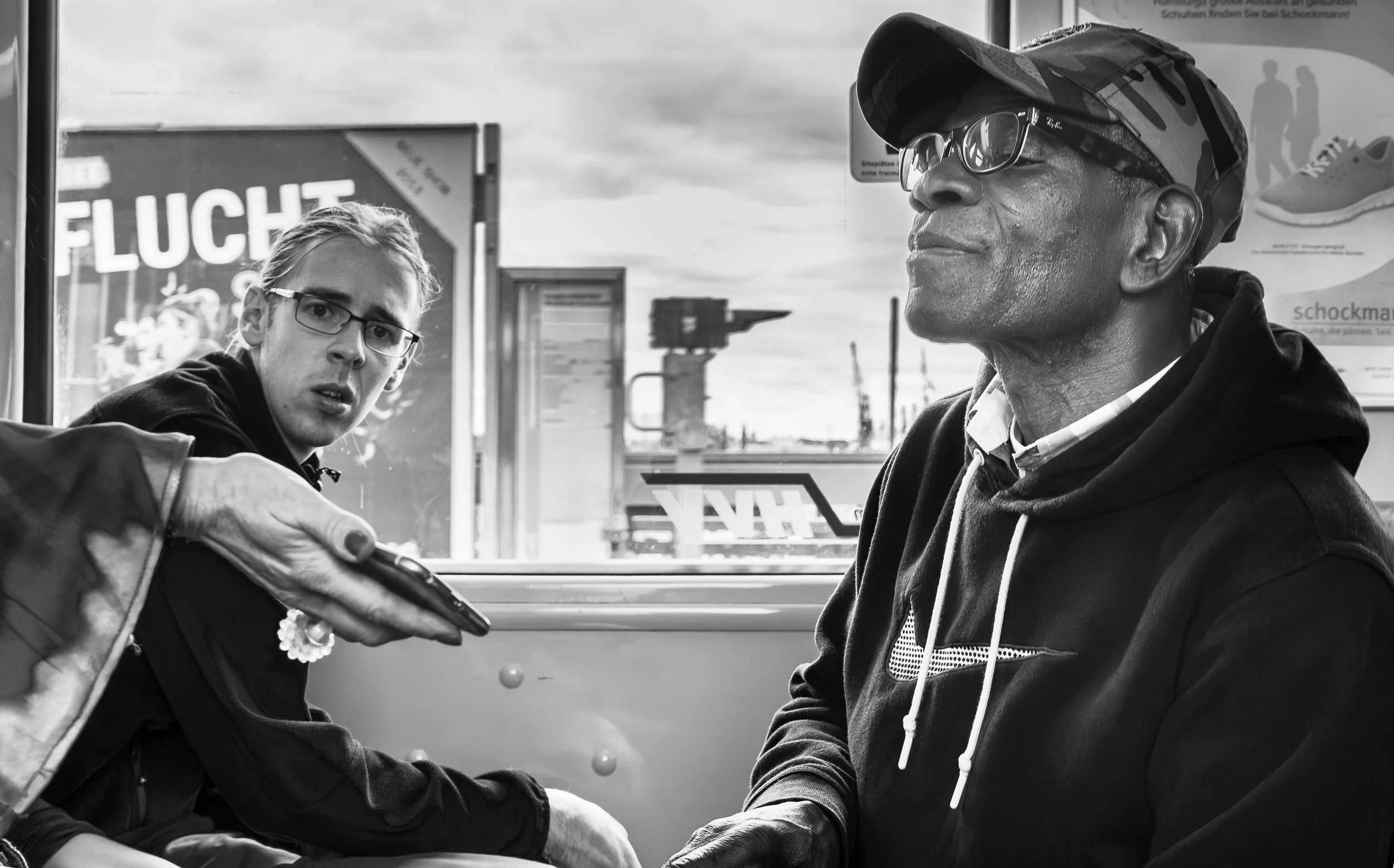 Two men seated on a bus, one person reaching out with a device or pen towards the other.