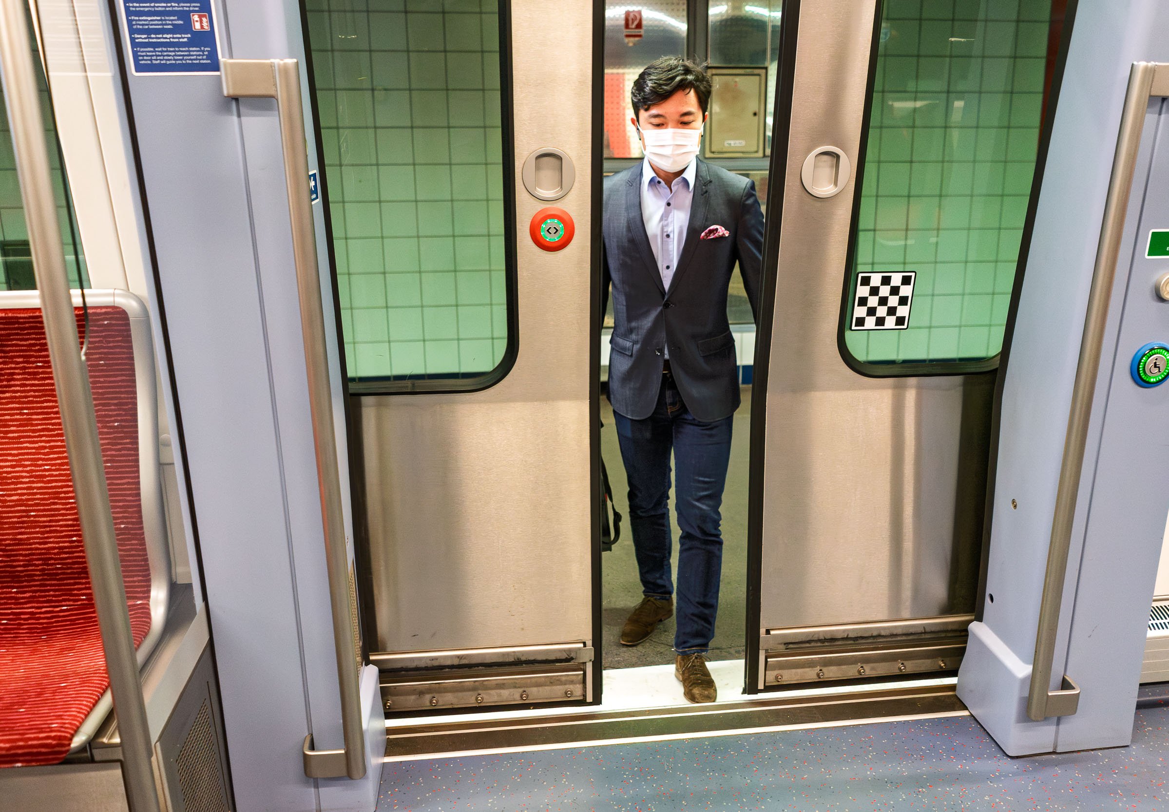 A man with dark hair wearing a suit and a face mask entering a subway train through open doors.