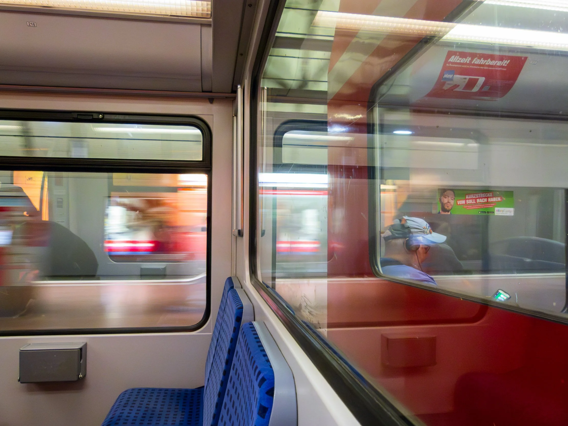 Inside a train, showing a blue seat and a large window with view of a blurred outside scene, and a reflection of a person with a cap in the glass.