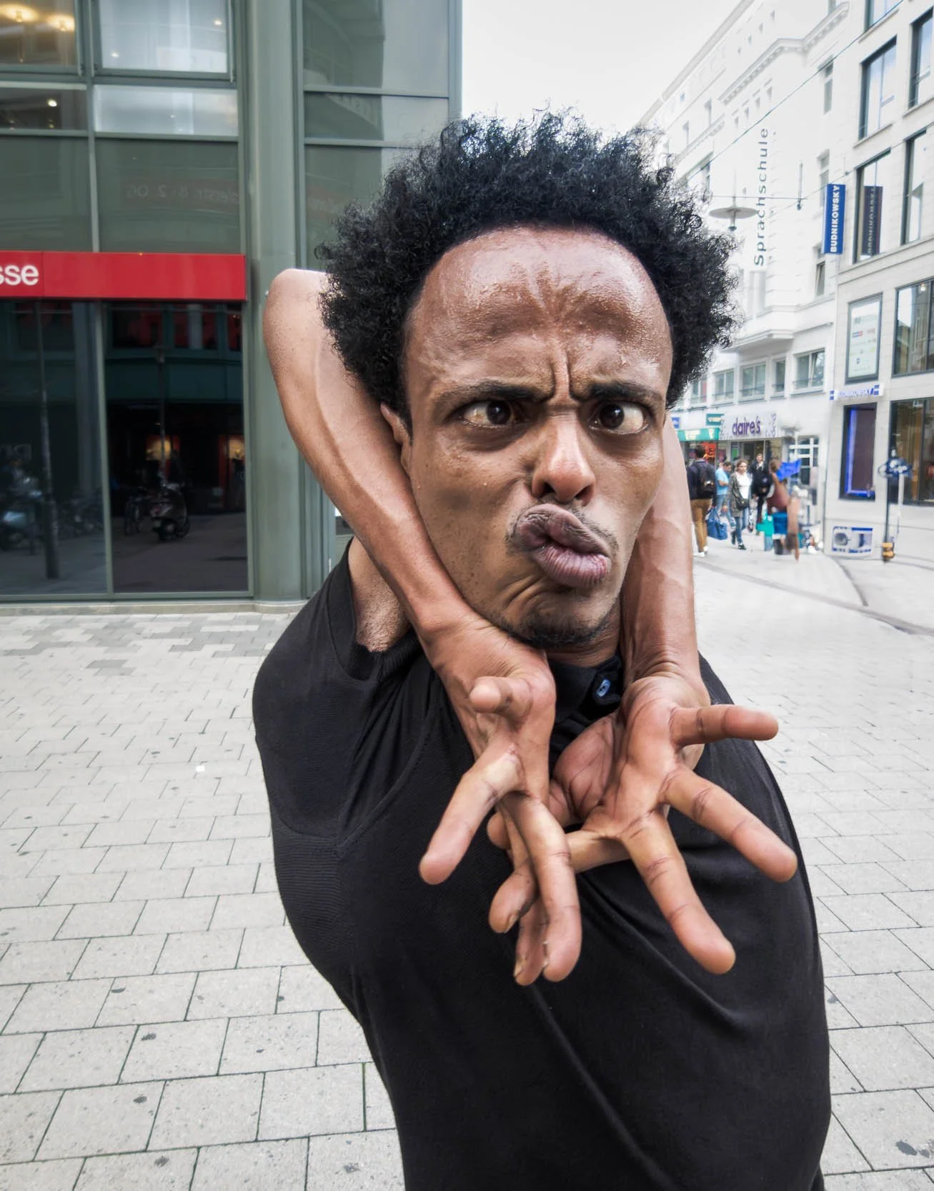 A man making a funny face with a furrowed brow and puckered lips, holding his head with one hand and pointing with the other, standing on a city street.