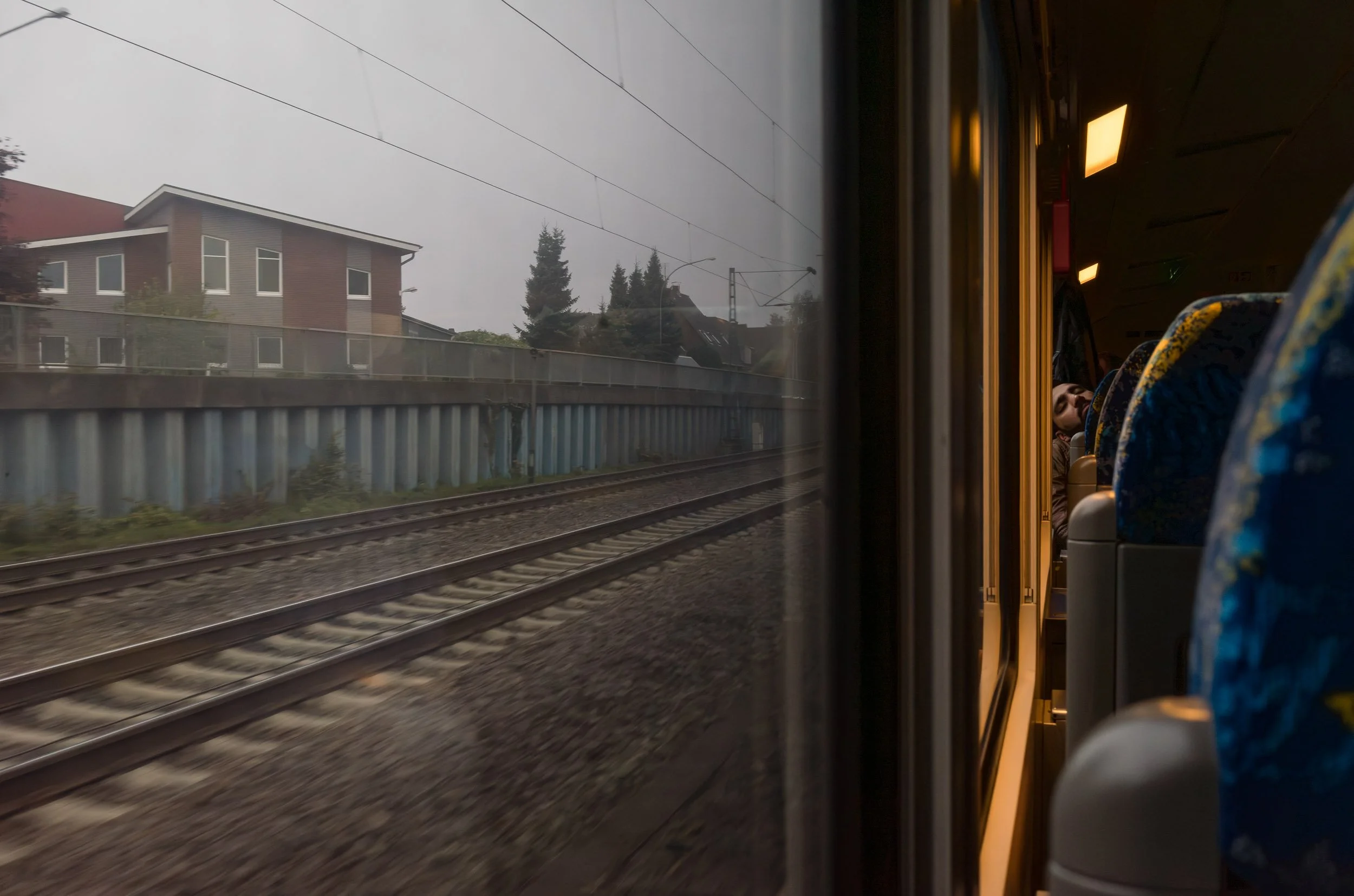 View from a moving train window showing railroad tracks, a fence, and residential buildings outside; inside the train, a woman is visible sitting and resting against the window.