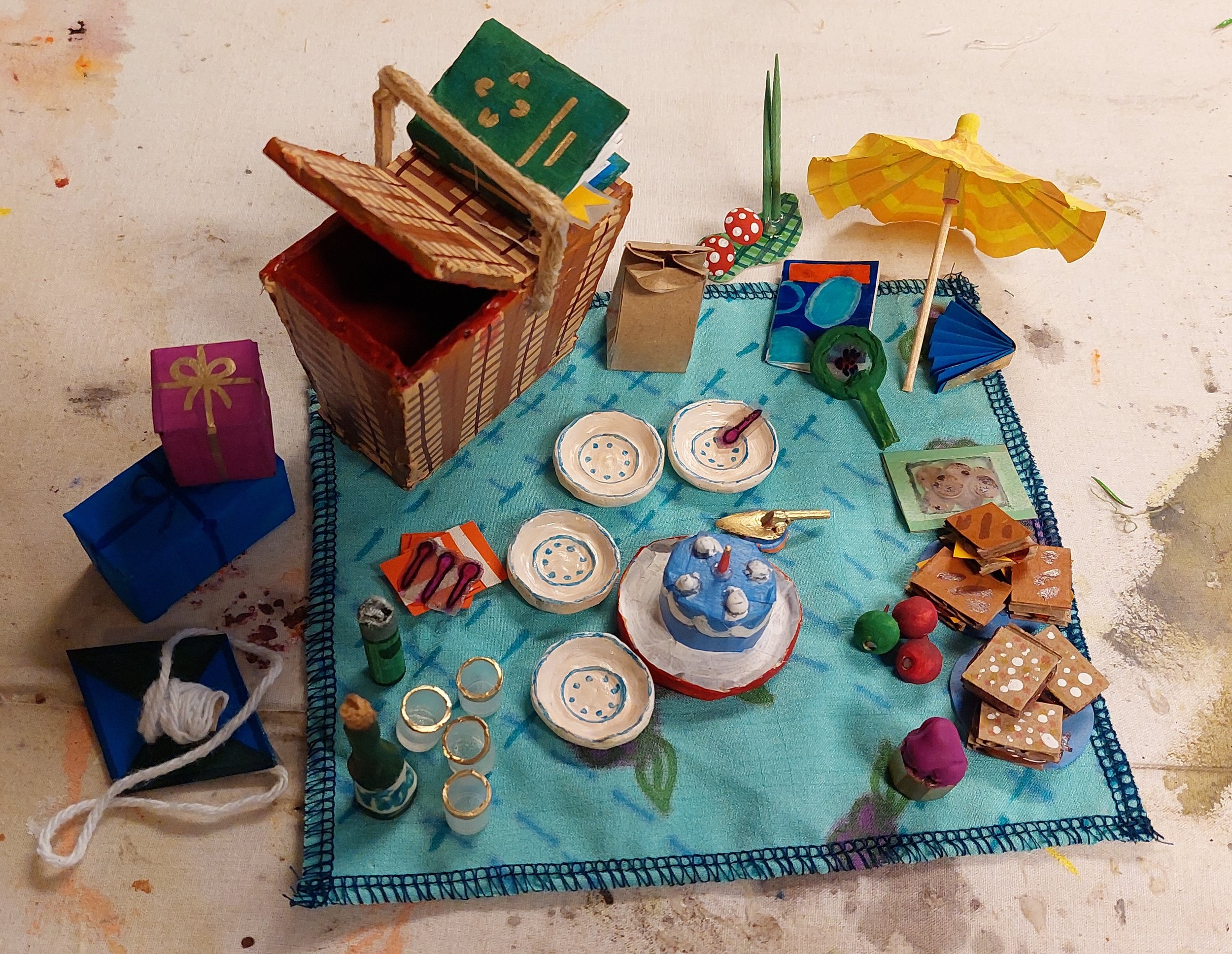 Miniature picnic set. Made with a variety of scrap and recycled materials including but not limited to paper, fabric, air dry clay, beads, cardboard, string, epoxy glue, tape, and a drink umbrella, for scale.