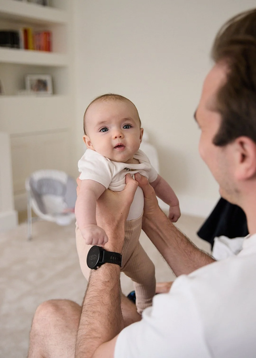 A man holding a baby up in a room with bookshelves and a chair in the background.