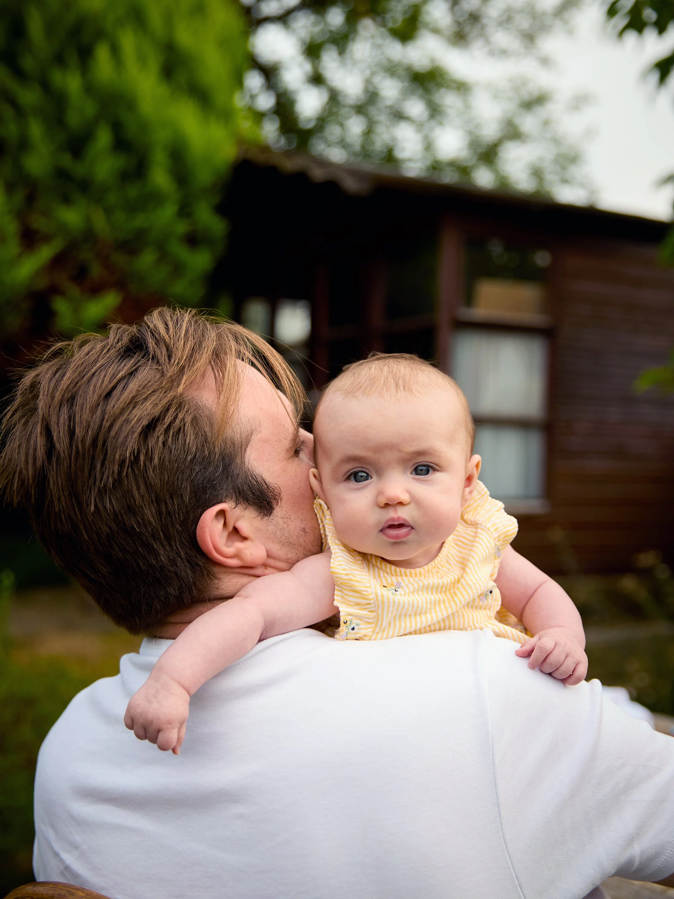 A man holding a baby girl outdoors with a house and trees in the background.