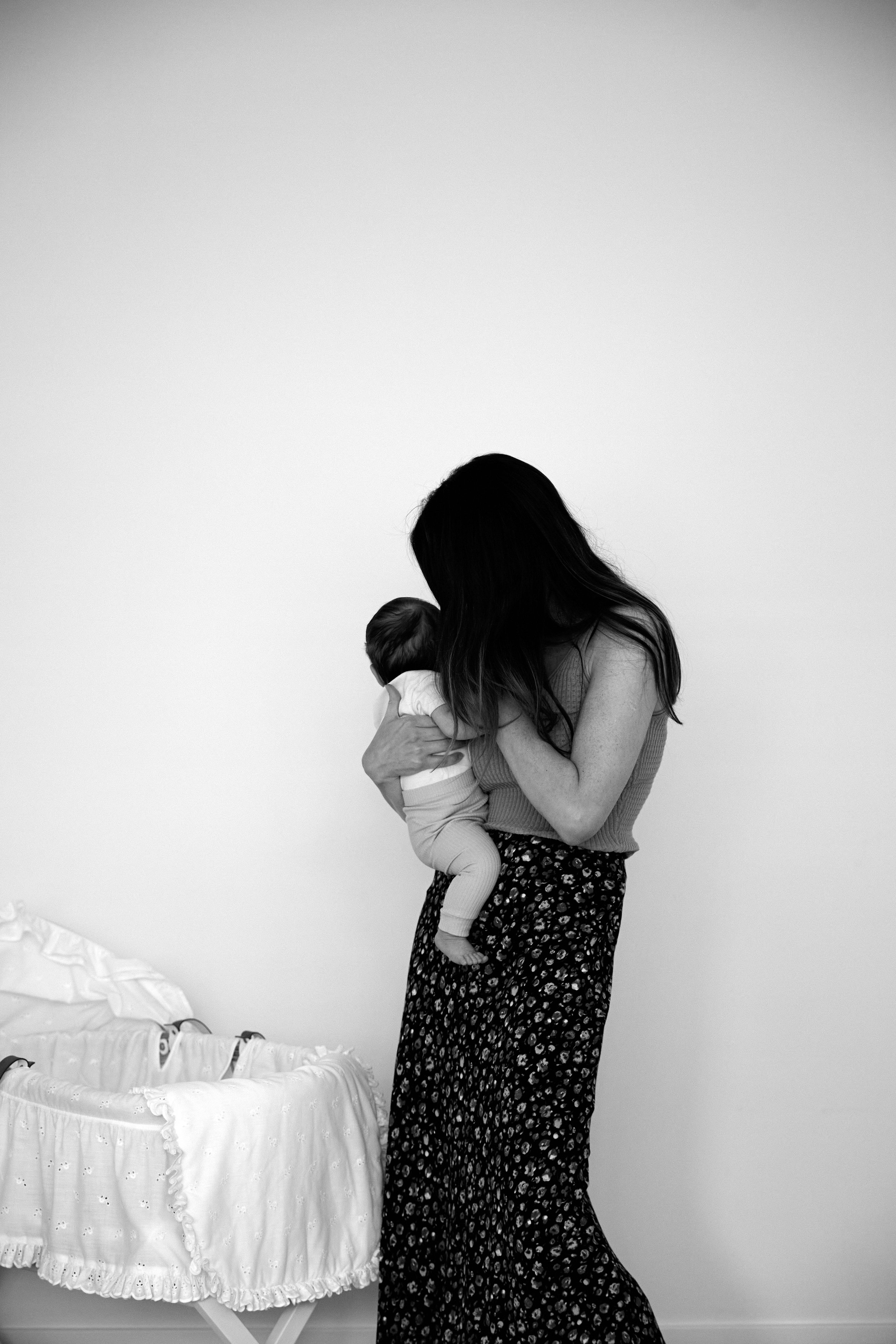 A woman holding a baby near a crib with a white blanket, inside a plain room.