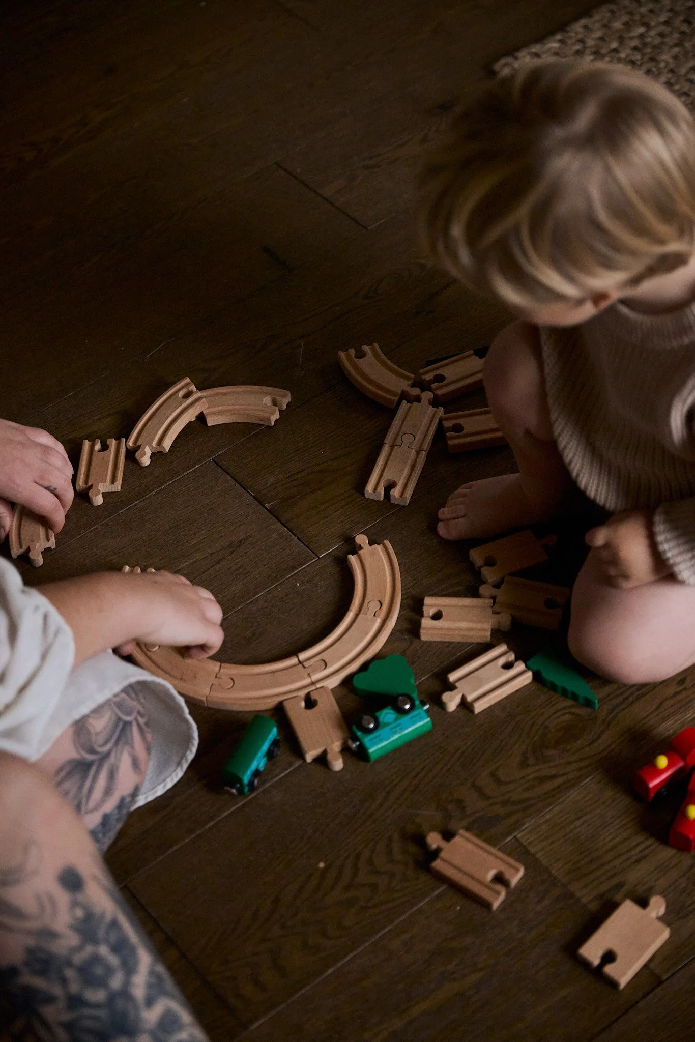Two children and an adult assembling a wooden train set on a dark wood floor, with train tracks and colorful trains.