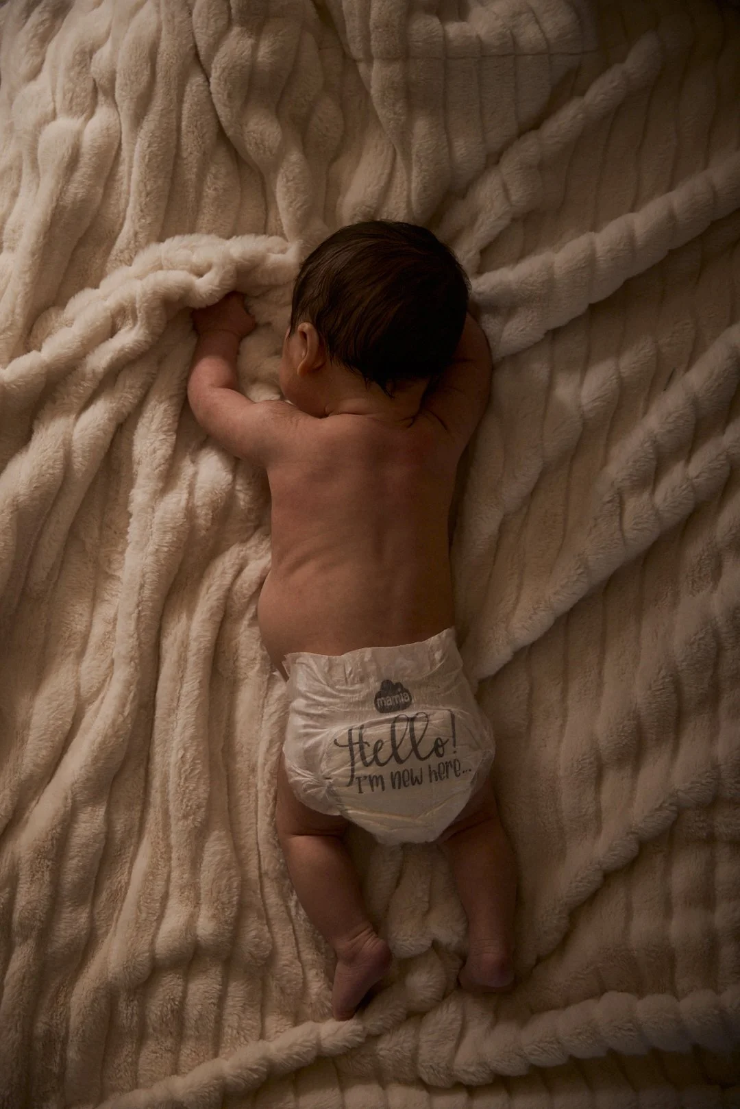 A sleeping baby lying on a plush, cream-colored blanket, wearing a diaper that says 'Hello! I'm new here...'