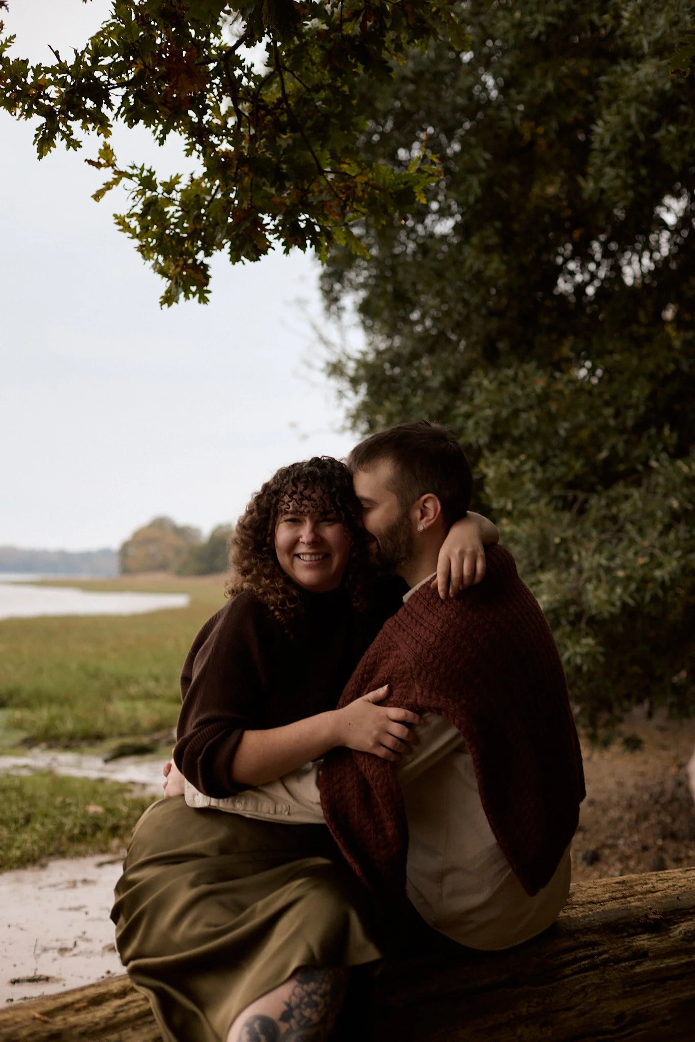 A couple sitting on a log outdoors, embracing and smiling, with trees and water in the background.