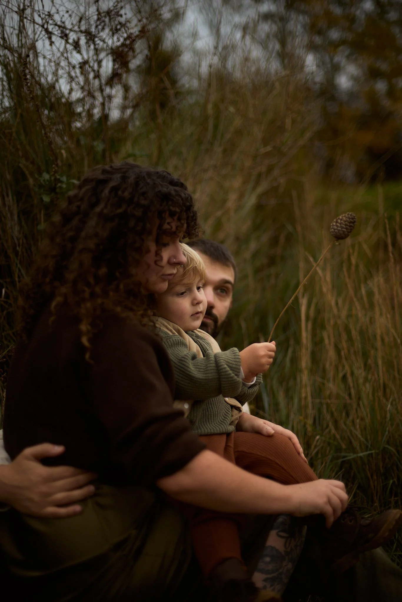 A woman, a man, and a young child sitting outdoors among tall grasses, the child holding a pinecone on a stick.