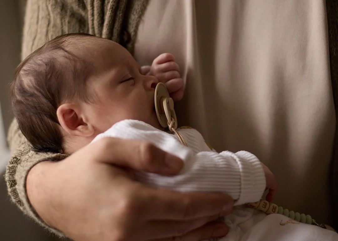A baby sleeping with a pacifier in its mouth, being held gently in an adult's arms.
