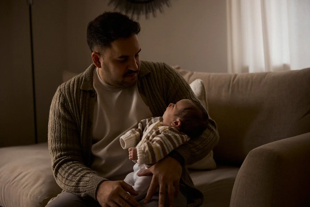 A man holding a baby in a cozy living room, looking at the baby with affection.