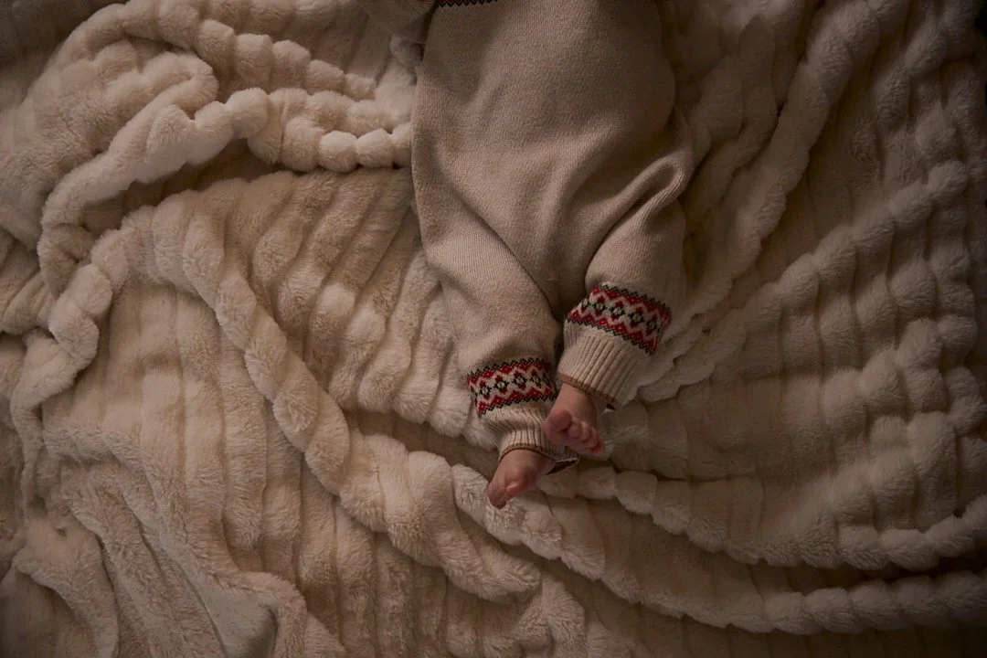 A newborn baby lying on a plush, beige faux fur blanket, wearing a cozy beige sweater with patterned cuffs. Only their feet and part of the sleeve are visible.