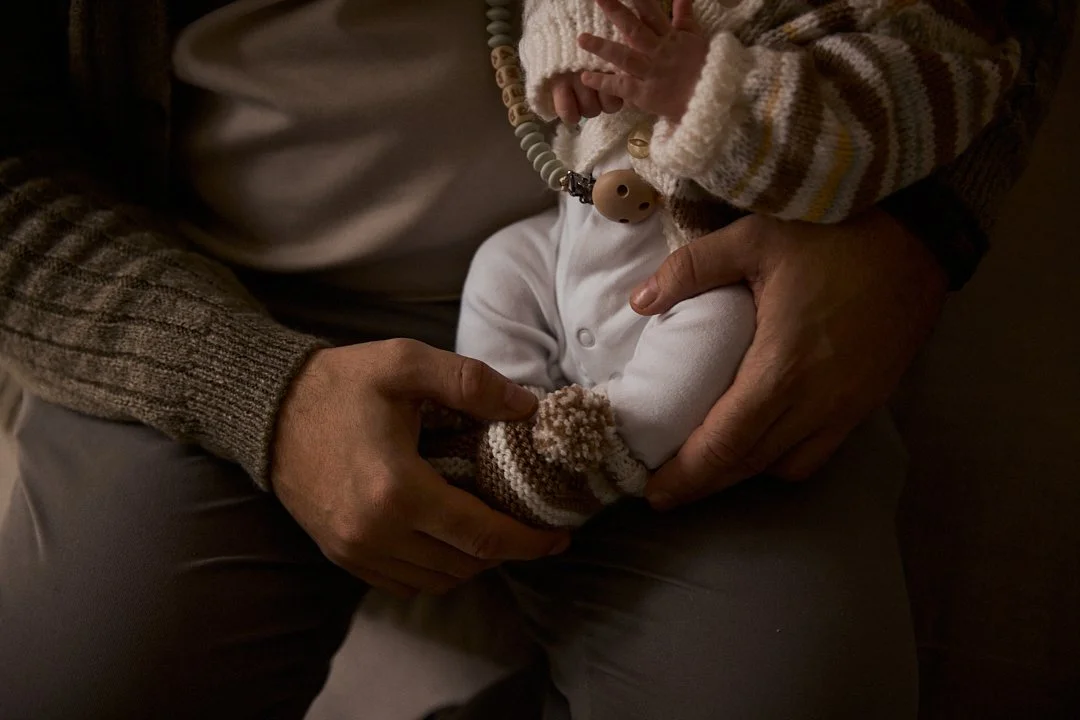 An adult holding a baby in their lap, with the adult's hands gently supporting the baby's legs and body.