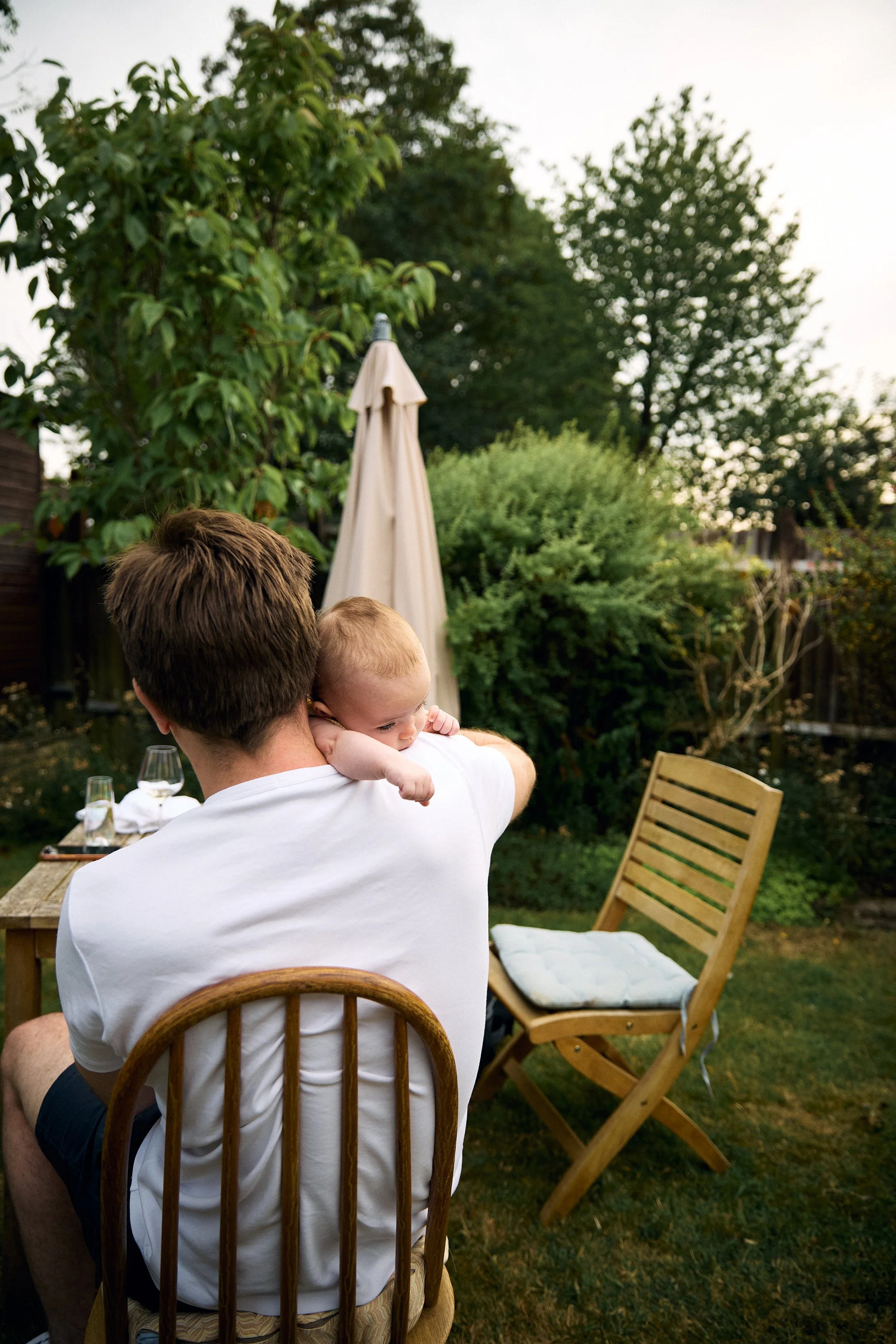 A man is sitting on a wooden chair in a backyard, holding a baby on his shoulder. The backyard has a table with glasses and a folded napkin, surrounded by trees and bushes, with a closed umbrella in the background.