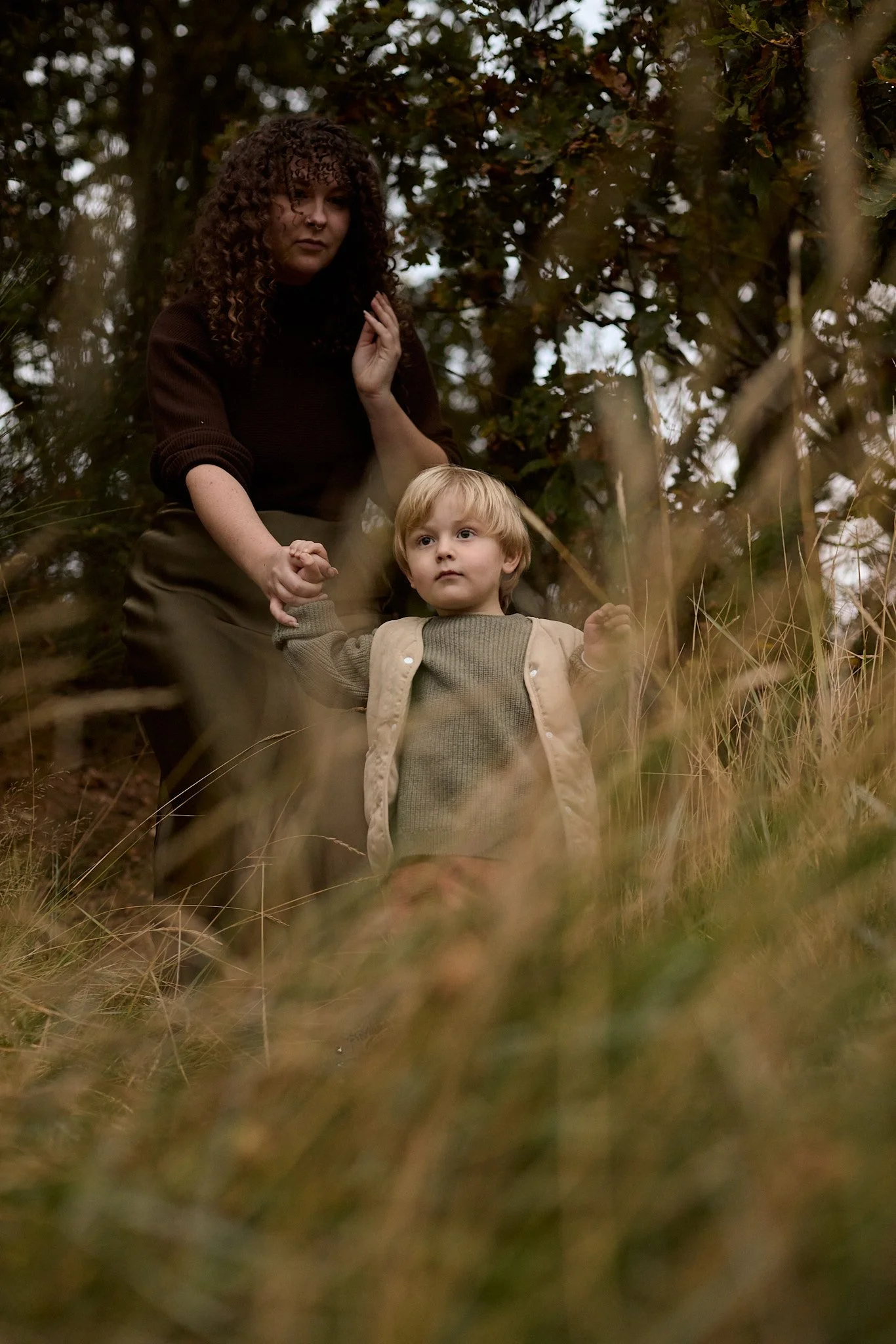 A woman with curly hair holding a young child's hand while standing in a woods with tall grass and leafy trees.