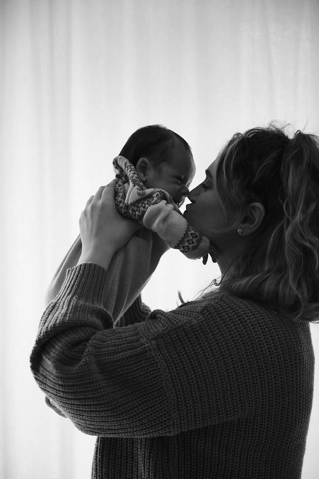 A woman lifts a young child, touching her nose to the child's nose, both with their eyes closed, against a plain, light background.