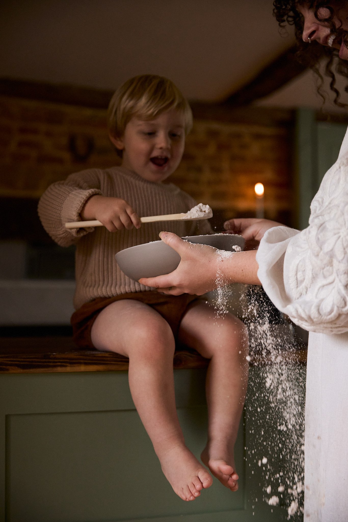 A young boy with blonde hair sitting on a kitchen counter, smiling as a woman, likely his mother, helps him sprinkle flour into a bowl.