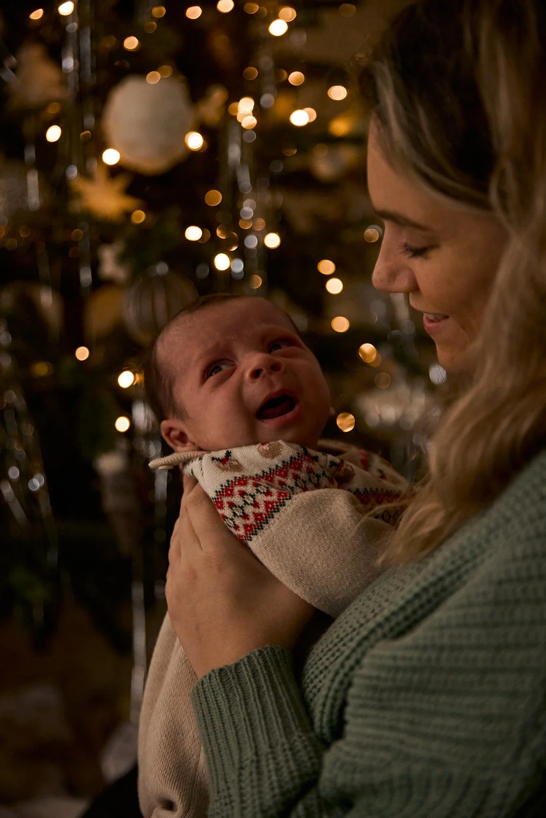 A woman holding a crying baby in front of a decorated Christmas tree with lights.