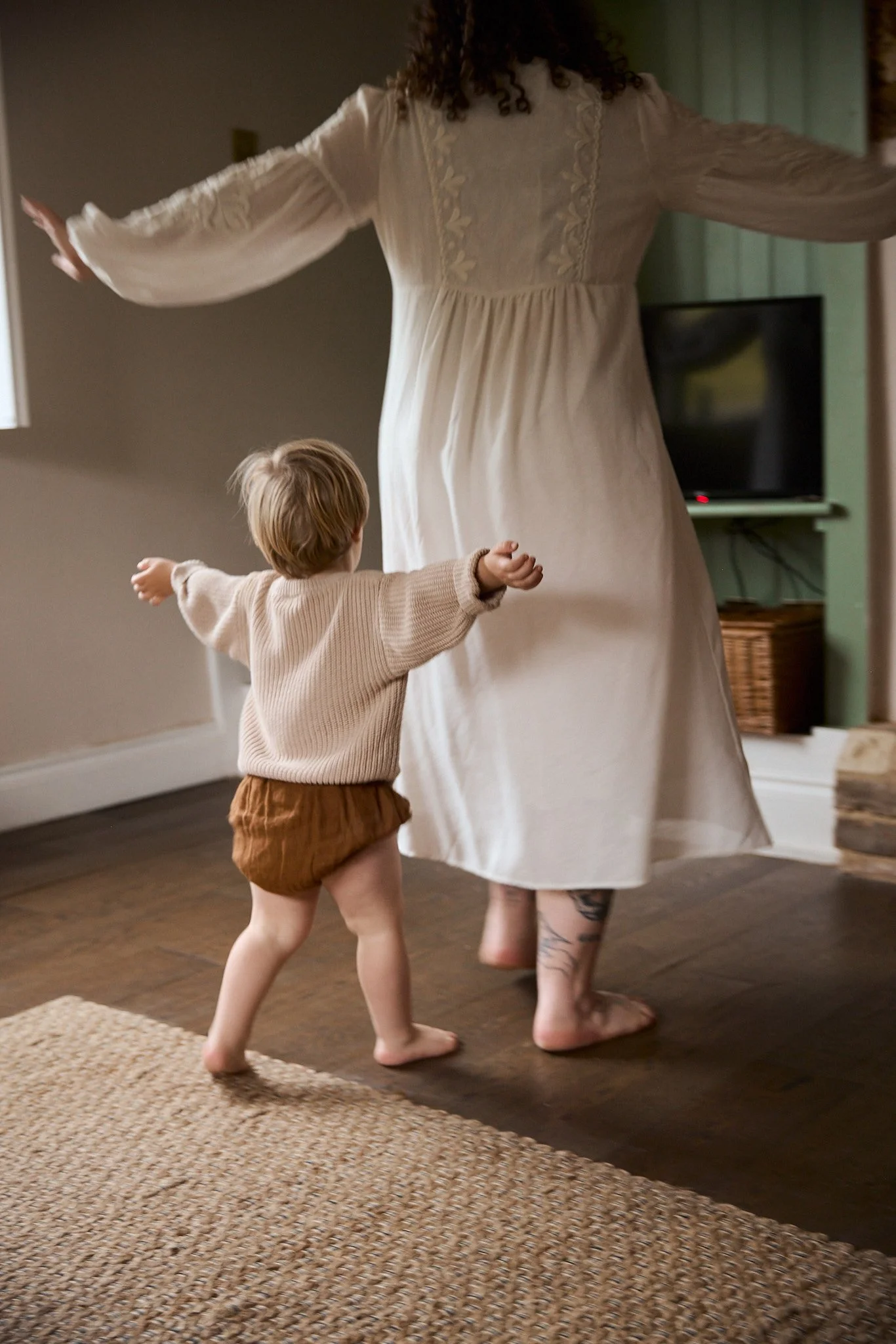 A young child and an adult dancing in a living room. The child wears a beige sweater and brown shorts, and the adult wears a long cream-colored dress with embroidery. The adult's face is not visible, and the child appears to be happily moving with ar