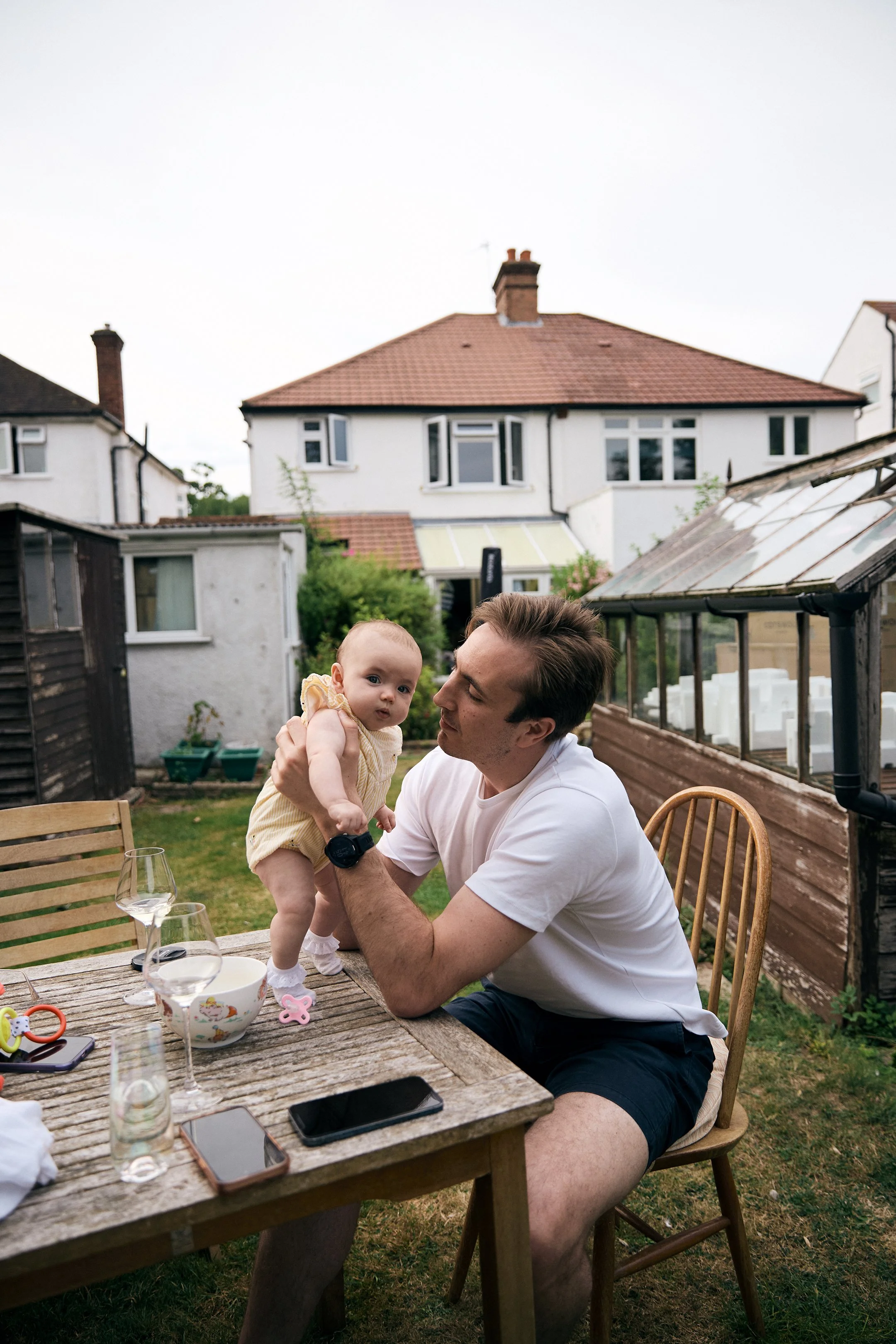 A man sitting at a wooden outdoor table holding a baby girl. There are empty wine glasses, a bowl, and a smartphone on the table, with a house and garden in the background.