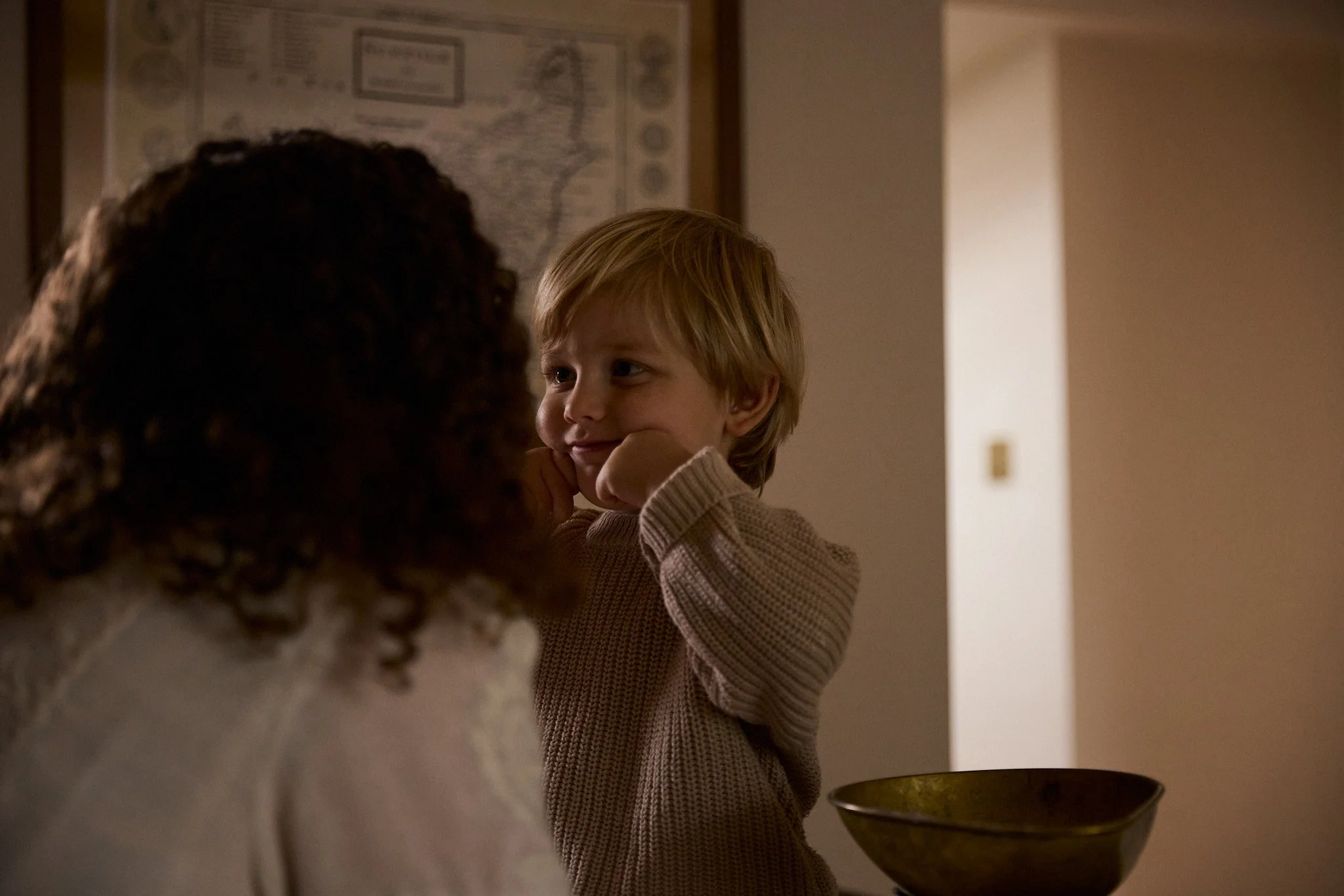 Young boy with blond hair leaning on his hand, smiling at a woman with curly hair, in a cozy indoor setting with a wall map in the background.