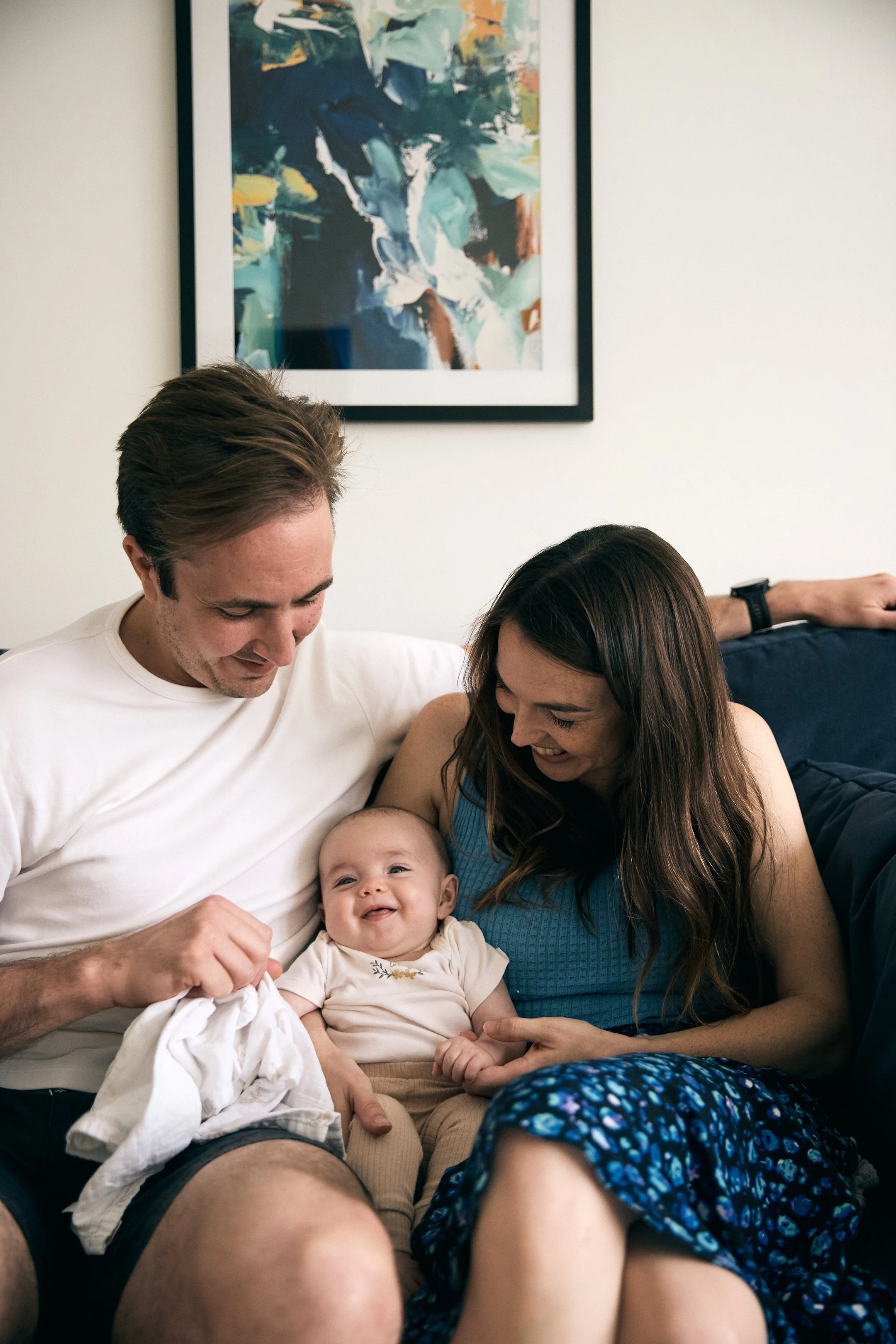A happy family of three, including a baby, sitting together on a couch and smiling at each other.