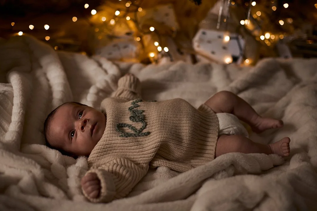 A baby lying on a plush blanket under a decorated Christmas tree with warm lights.