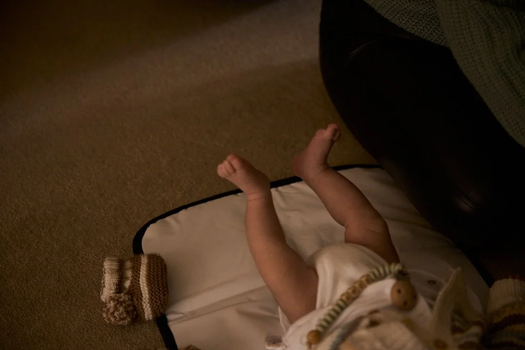 A baby lying upside down on a changing pad on a carpeted floor, with legs raised in the air and a wrapped pacifier around the neck.