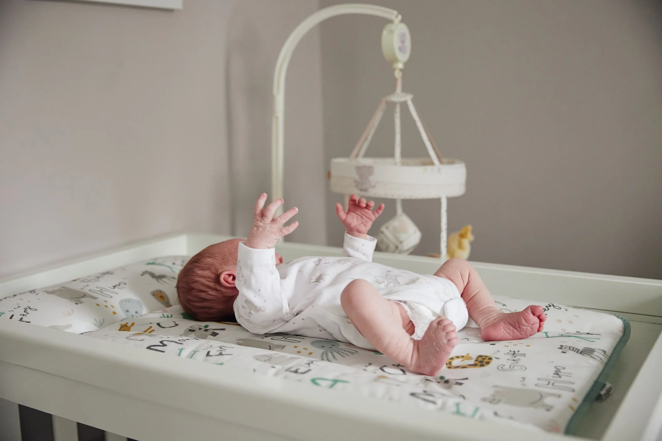 A newborn baby lying on a changing table in a nursery, with arms raised and eyes closed, surrounded by soft, neutral-colored decor and stuffed animals.
