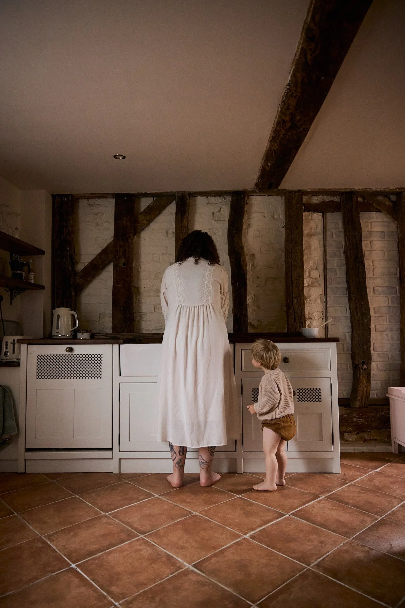 A woman and a young child standing in a rustic kitchen, facing the wall with exposed wooden beams. The woman, with curly hair and tattoos on her legs, is wearing a long white dress, and the child, with short blonde hair, is wearing a beige sweater an