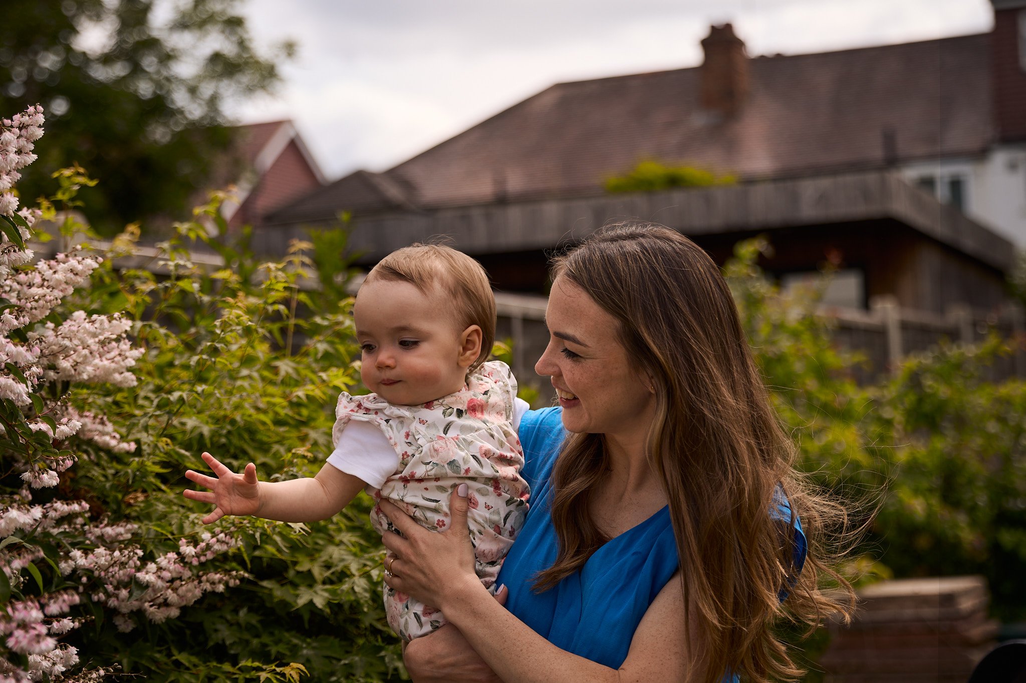 A woman holding a young child outdoors near pink flowering bushes, smiling, with houses in the background.