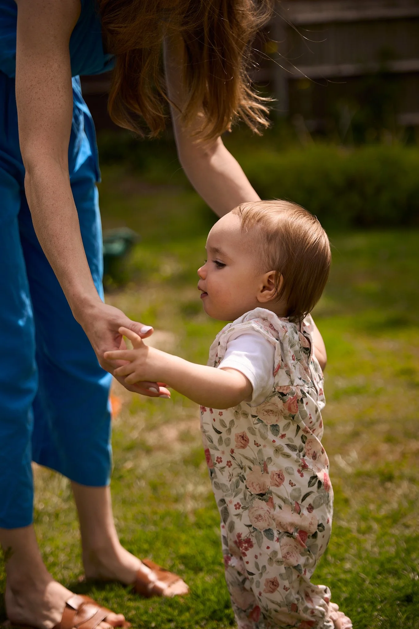 A young girl holding hands with a woman outdoors on grass, with a wooden fence and plants in the background.