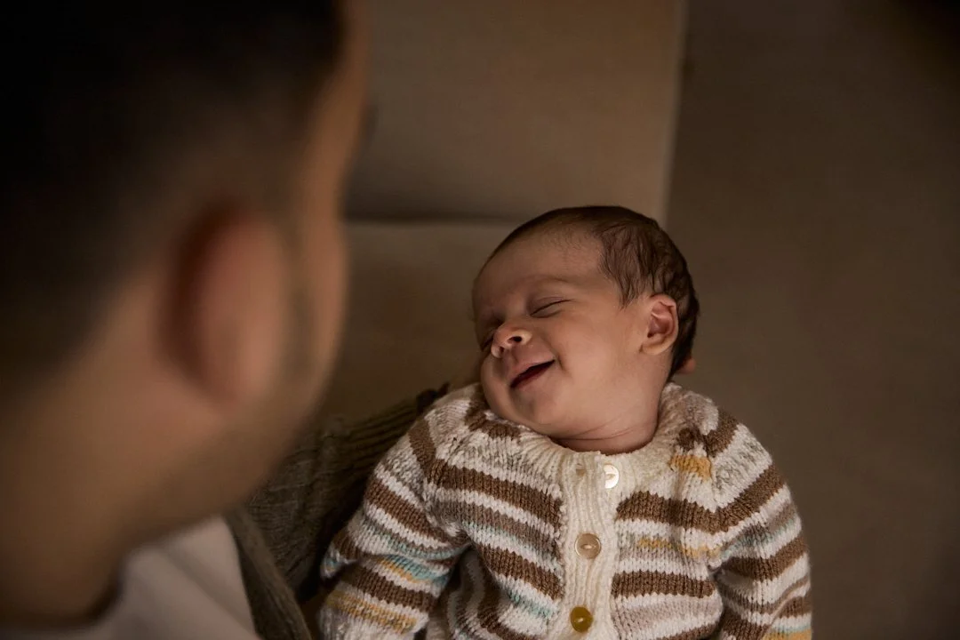 A man looks at a smiling baby lying on a beige couch, wearing a striped sweater.
