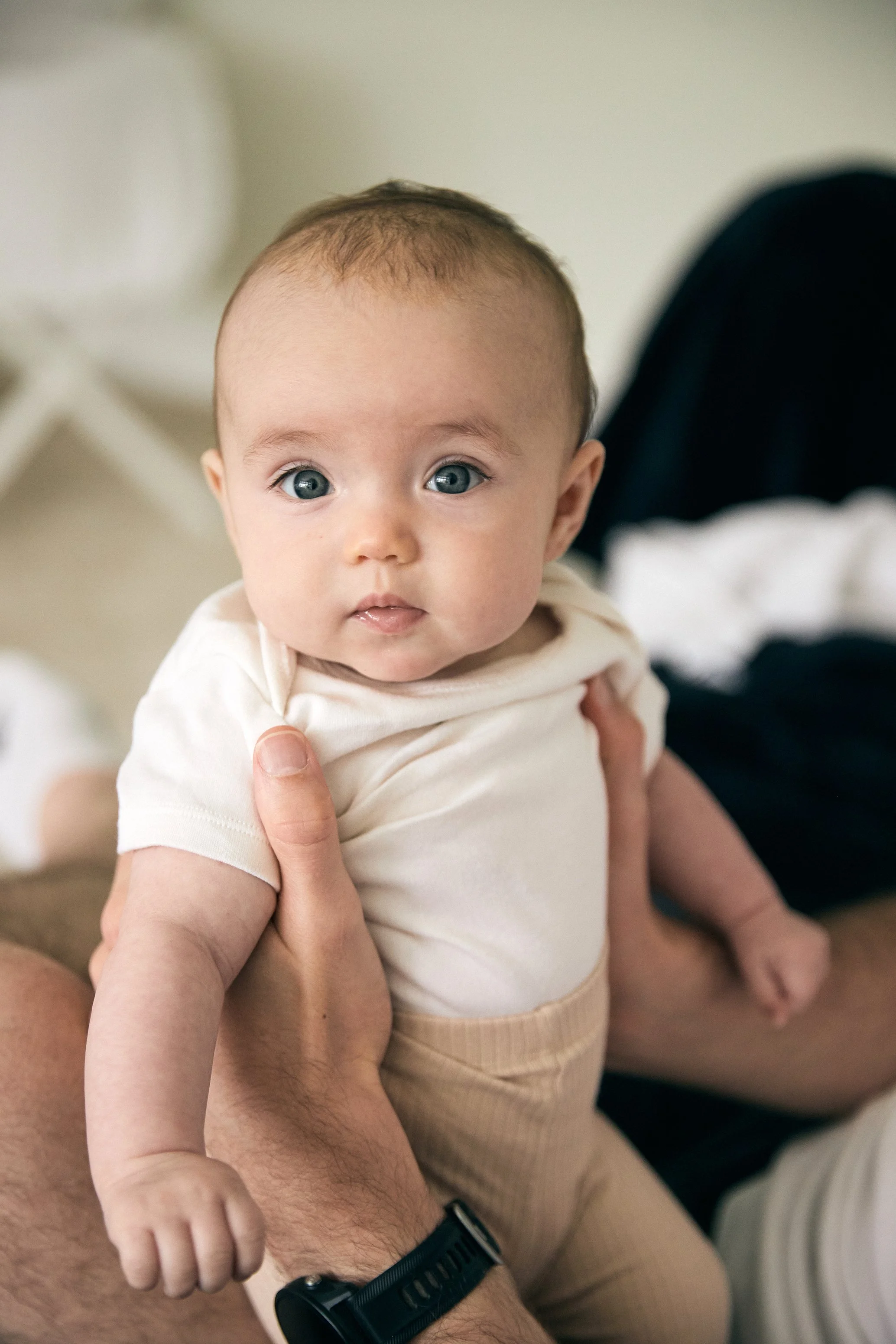 A baby with blue eyes, light brown hair, wearing a cream-colored shirt and beige pants, being held up by an adult's hand.