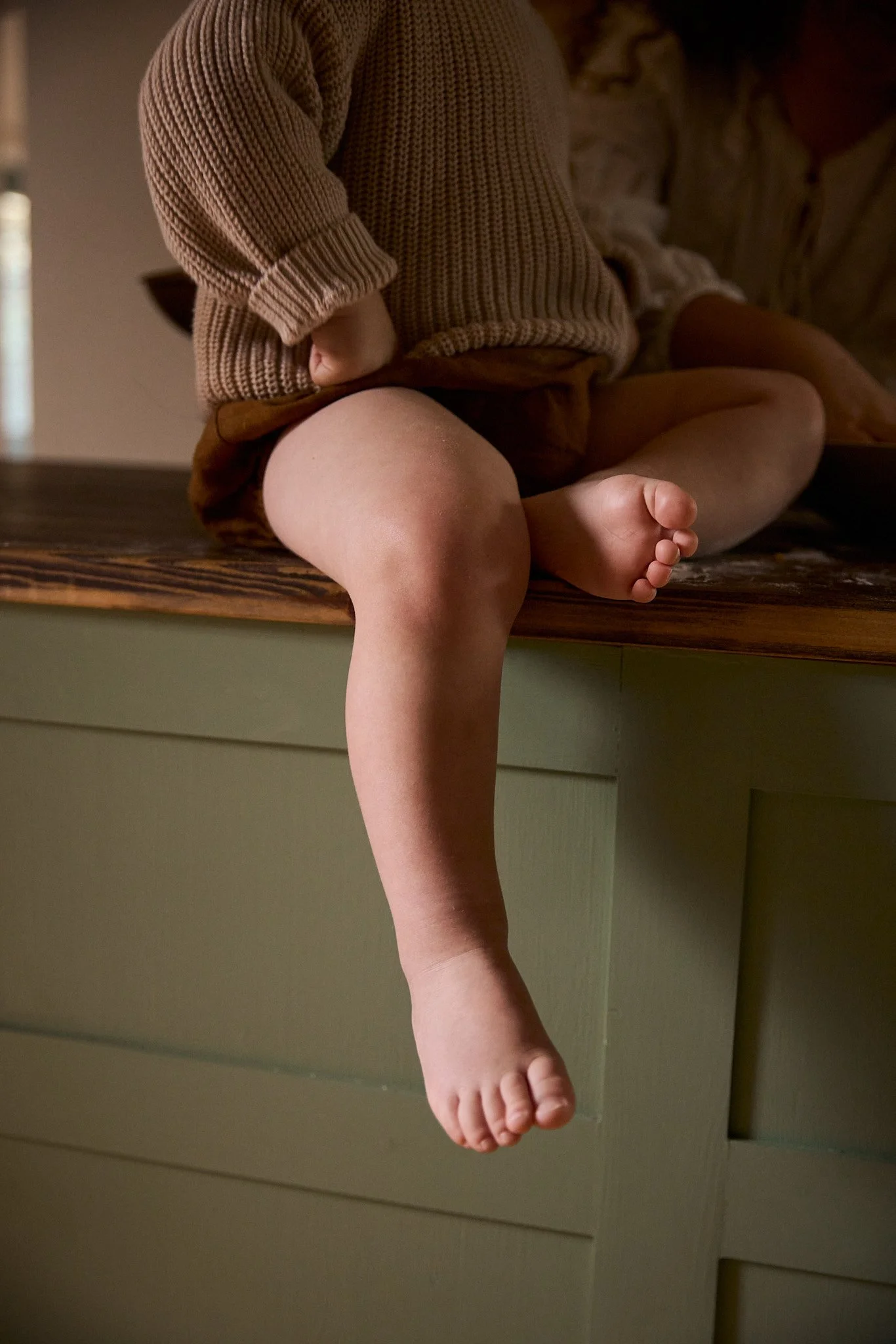 Close-up of a child sitting on a wooden surface with one leg hanging off, wearing a knitted sweater and brown shorts.