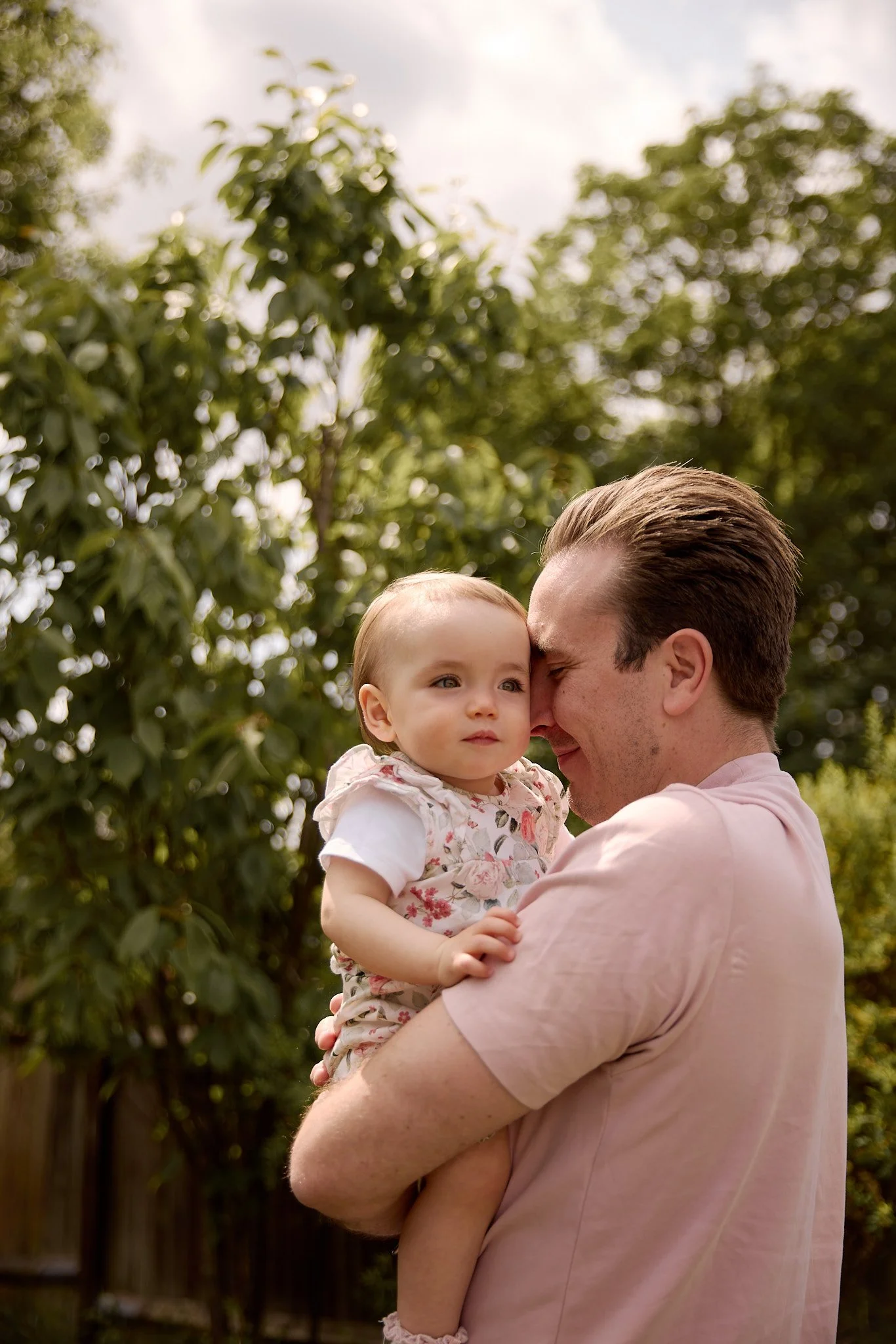 A man holding a young girl outdoors, with trees and a cloudy sky in the background.
