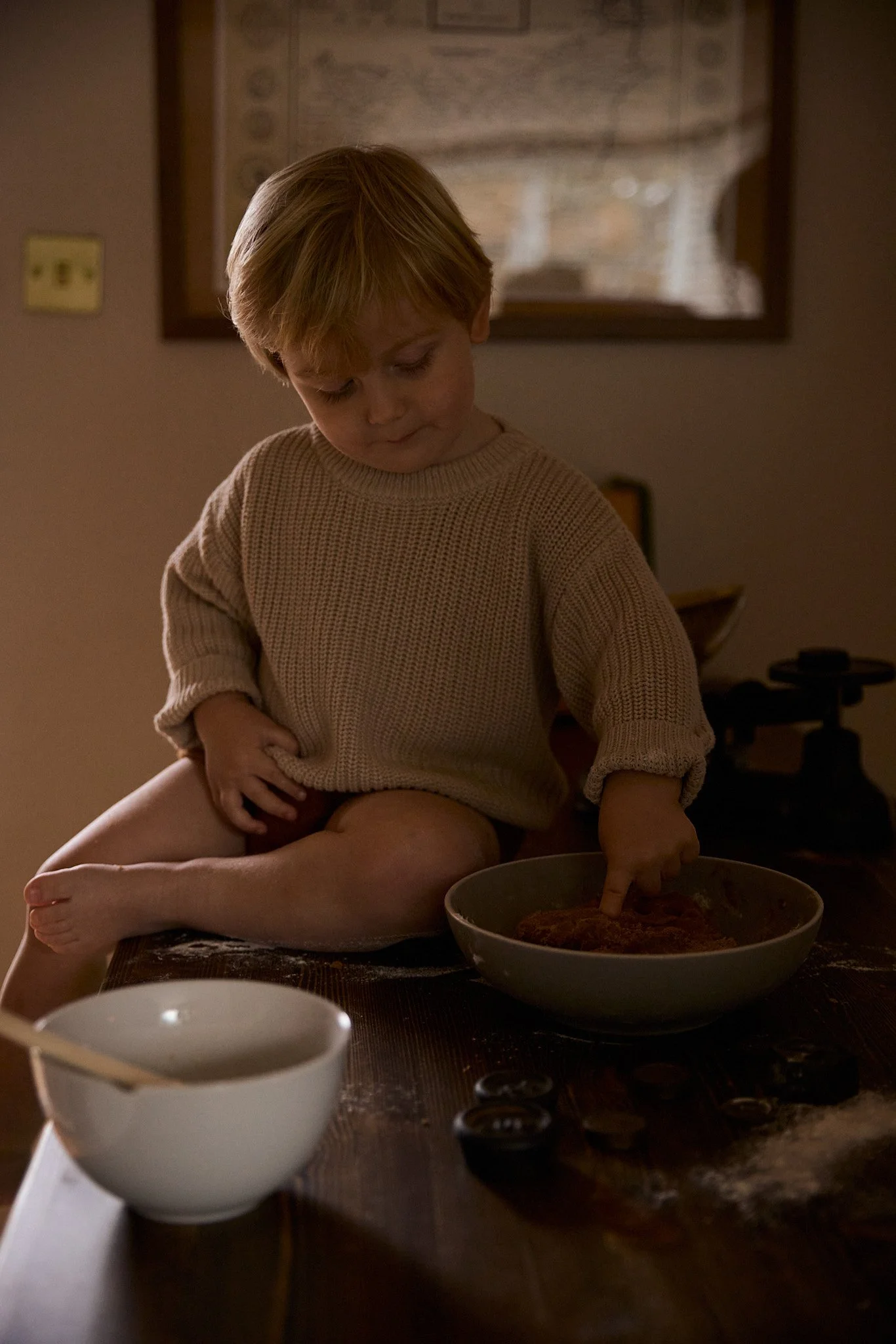 A young boy sitting on a wooden table, stirring or pressing into a bowl of dough or food, wearing a beige sweater, with a bowl and some small objects on the table, in a warmly lit room.