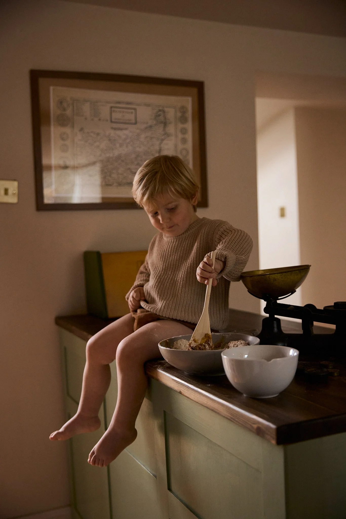 A young boy with blonde hair, wearing a beige sweater, is sitting on a kitchen counter with his legs dangling over the edge. He is stirring food in a large bowl with a white spatula. There are two bowls and a scale on the counter, and a map on the wa